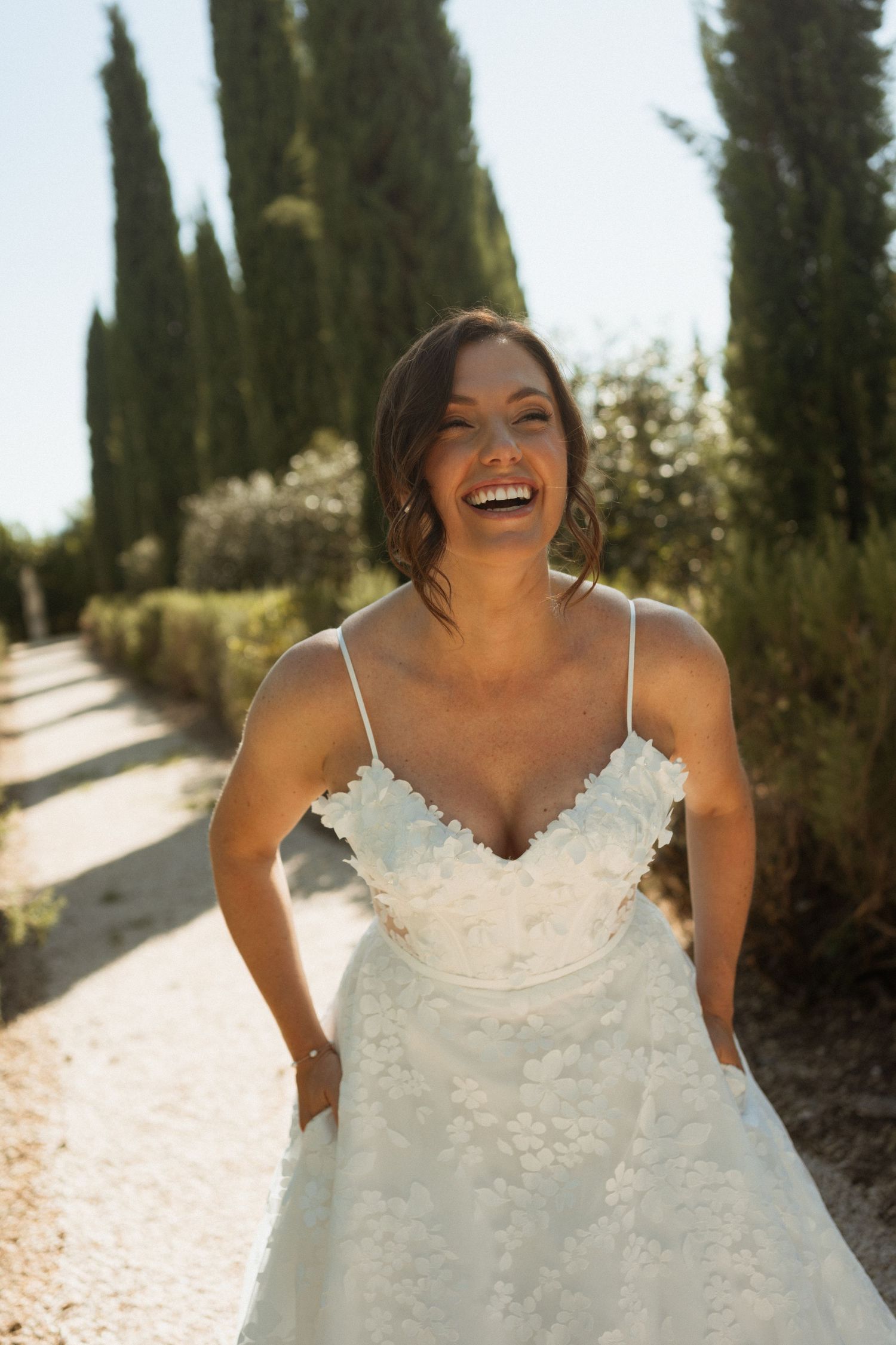 Joyful bride in a floral applique wedding gown laughing outdoors along a cypress-lined path.