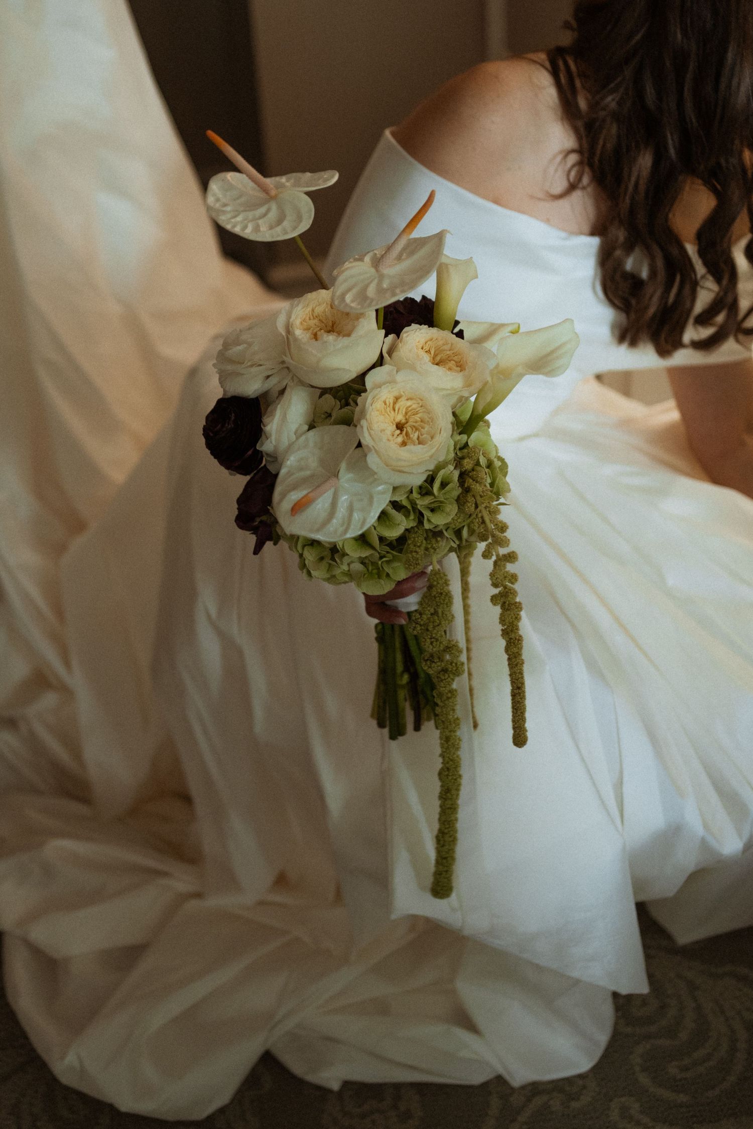 Bride in off-shoulder white satin gown holds bouquet of white anthuriums, roses, and green amaranthus.