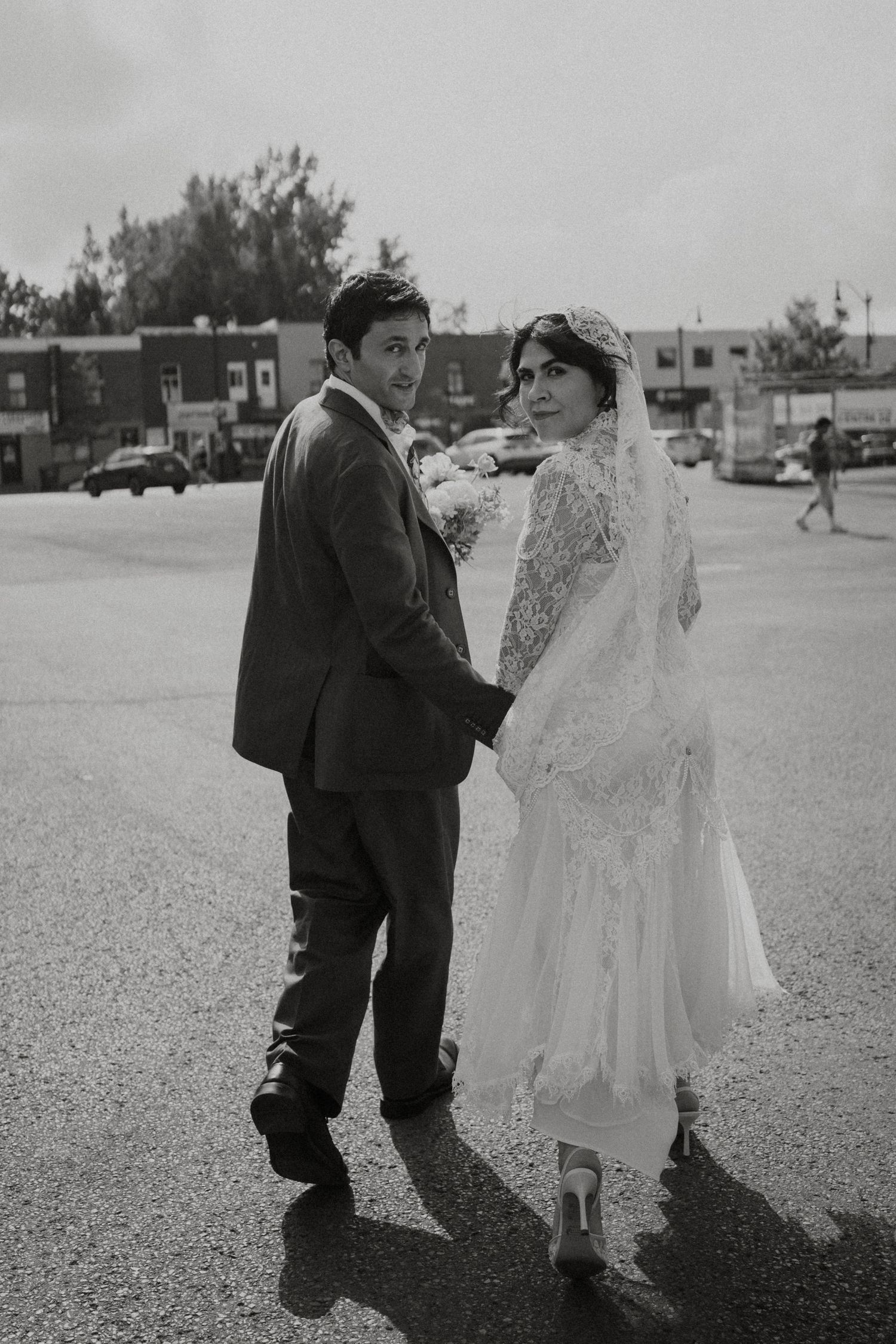Black and white photo of a bride and groom holding hands, walking in a parking lot, both looking back at the camera.