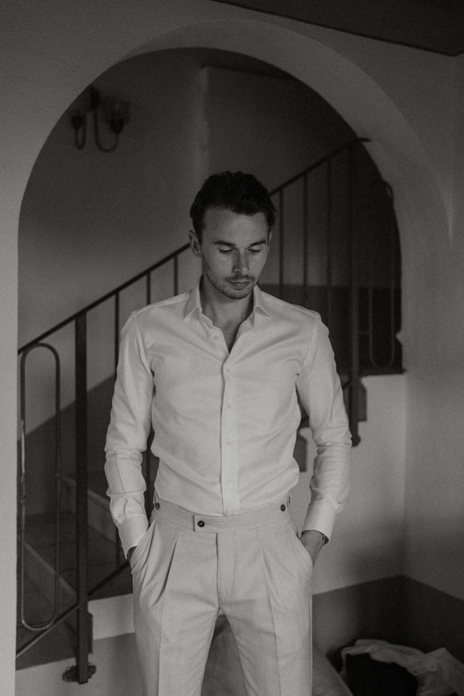 Black and white portrait of groom in white shirt standing under archway near staircase.