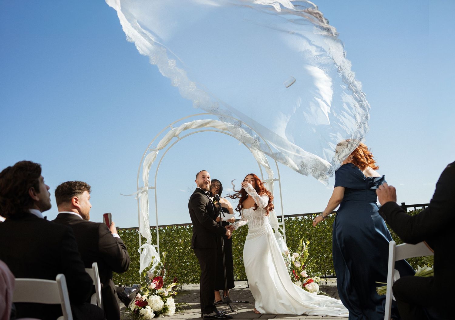 Outdoor wedding ceremony with bride and groom under floral arch as veil billows dramatically in wind against blue sky.