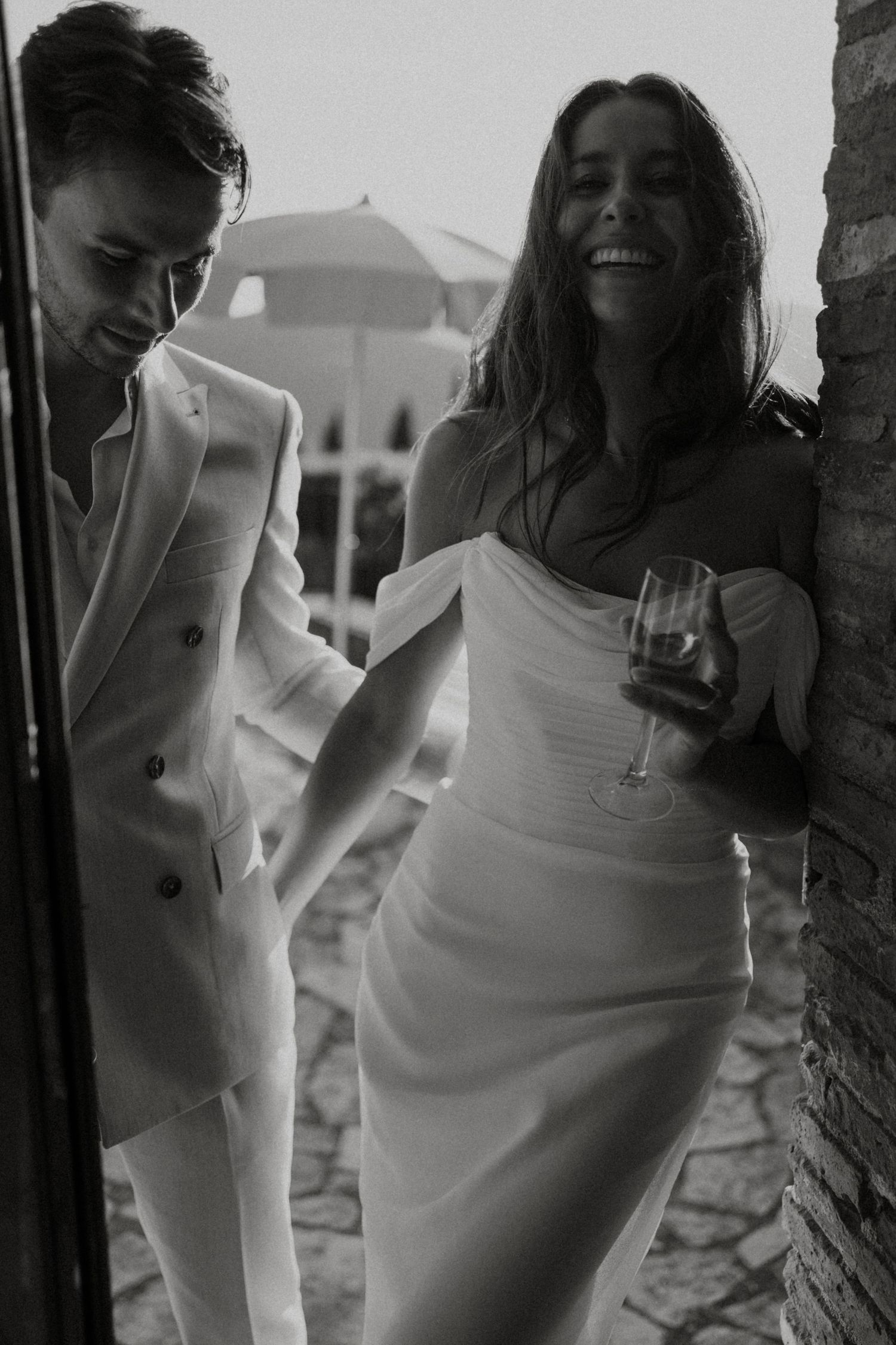 Bride in white off-shoulder gown laughing with groom in white suit in black and white wedding photo.
