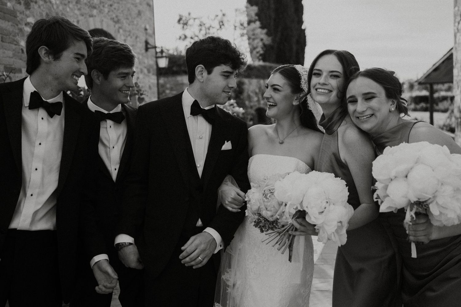 Black and white photo of bride, groom, and wedding party laughing together outdoors.