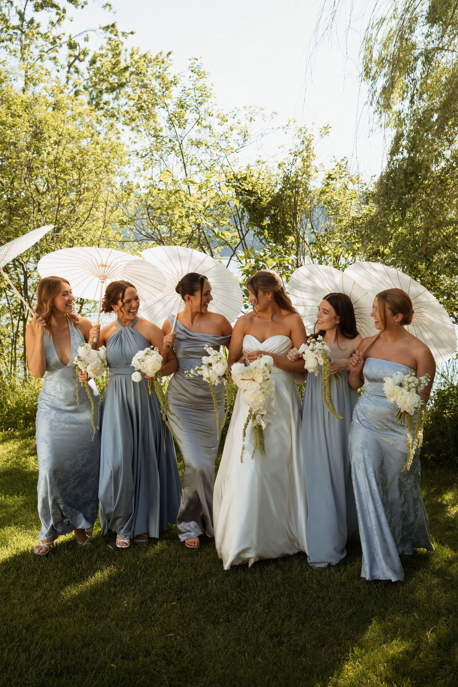 Bride with bridesmaids in blue satin gowns holding white parasols and bouquets in sunny garden setting.