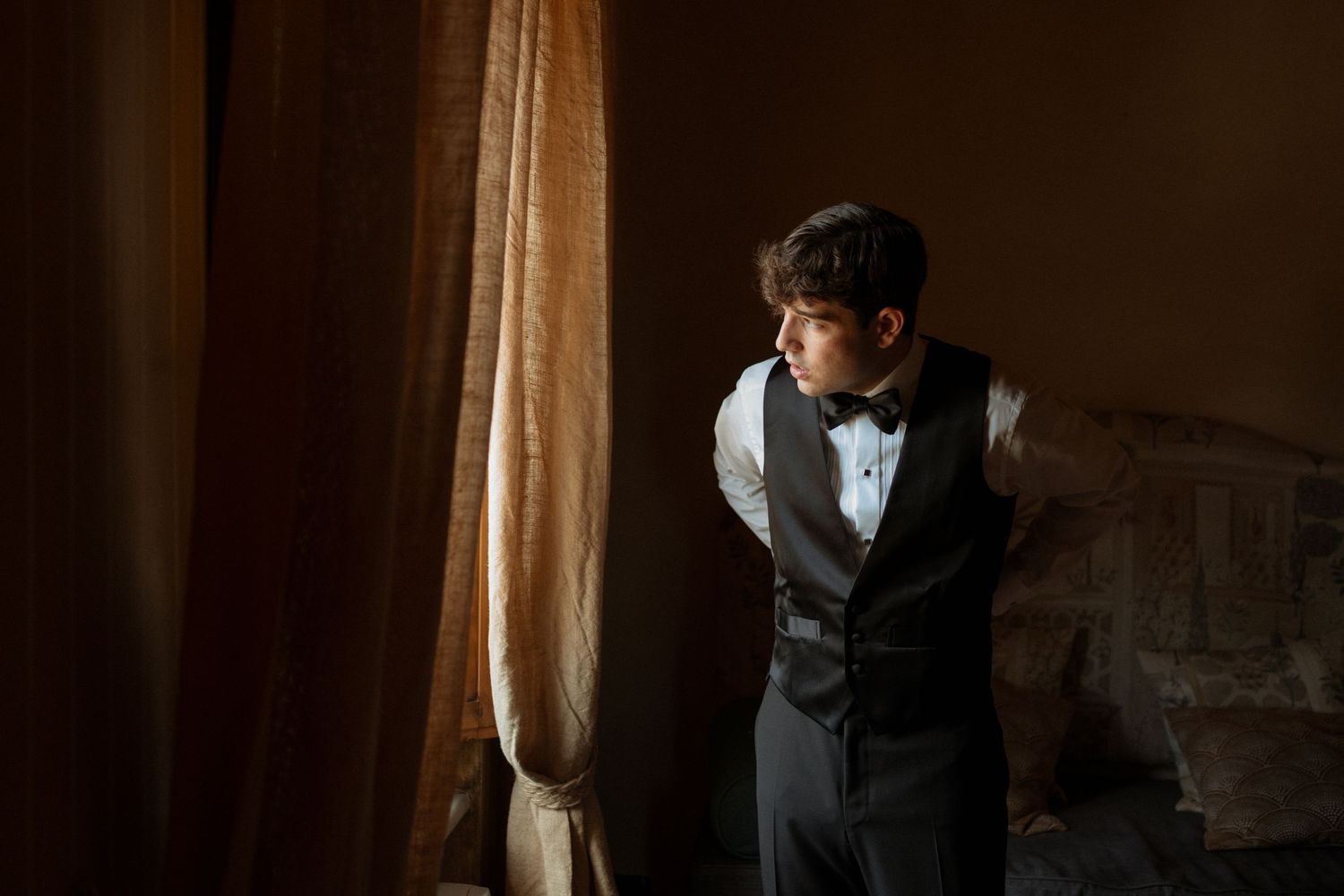 Groom in black tuxedo vest and bow tie standing by draped curtains in moody indoor lighting.
