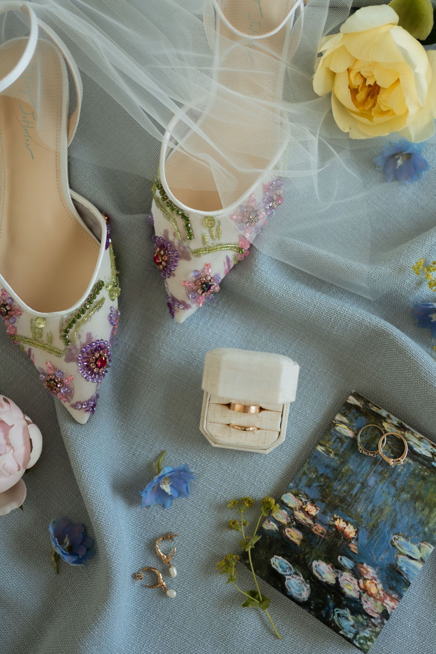 Elegant bridal flat shoes with floral embroidery beside wedding rings, earrings, and blue flowers on a soft blue fabric.