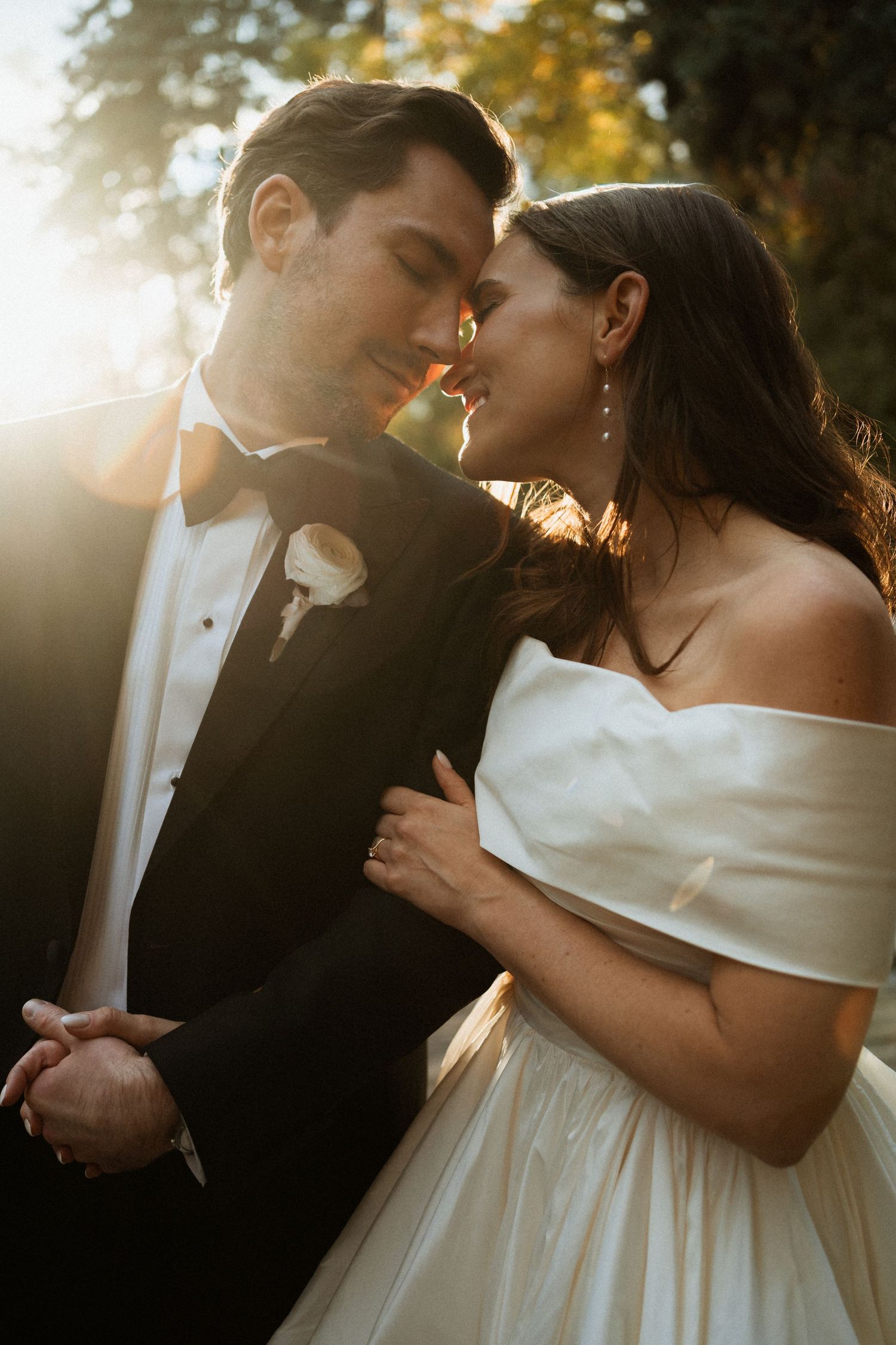 Bride and groom share intimate moment in golden sunset light, groom in black tuxedo, bride in off-shoulder gown.