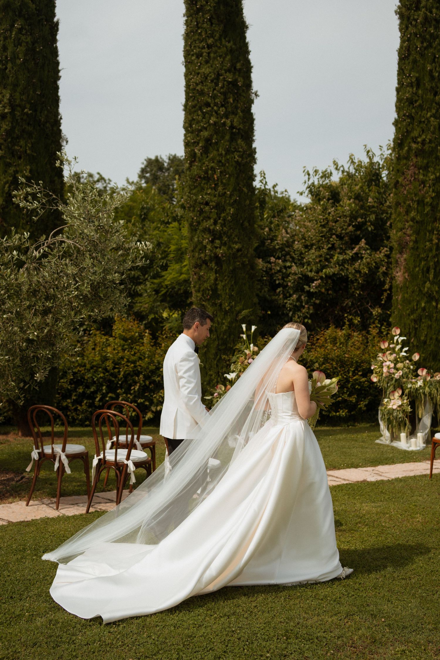 Bride and groom walking hand in hand through a lush garden with tall cypress trees at their wedding.