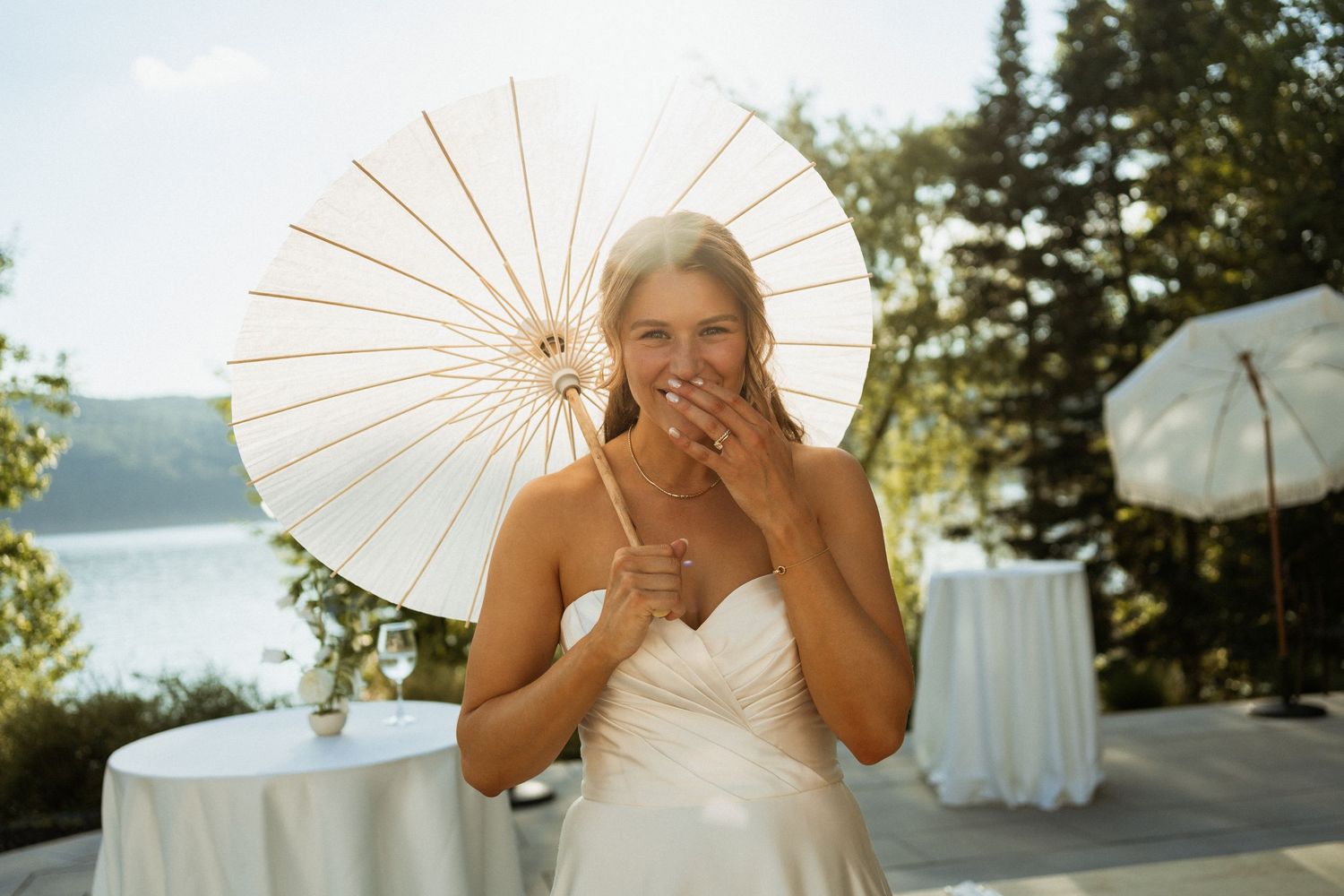 Smiling bride in white dress holds parasol at lakeside outdoor wedding reception on sunny day.