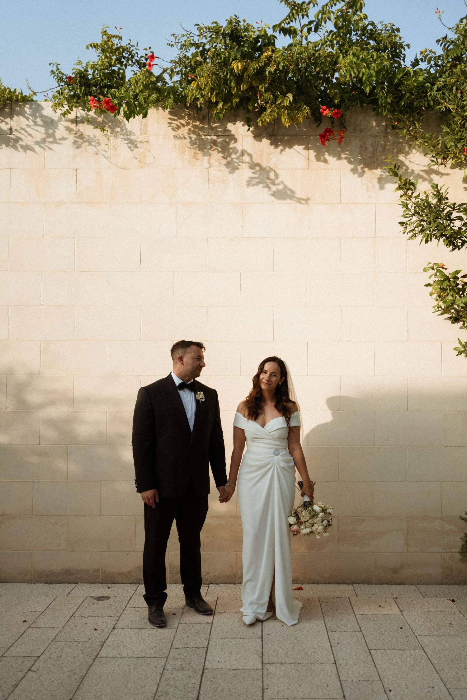 Bride and groom holding hands before sunlit stone wall with flowering vines at outdoor wedding venue.