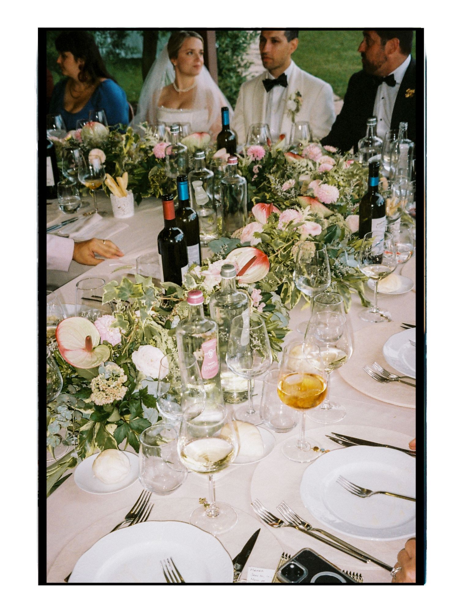 Elegant wedding reception table with pink floral centerpieces, glassware, and bride and groom seated in background.