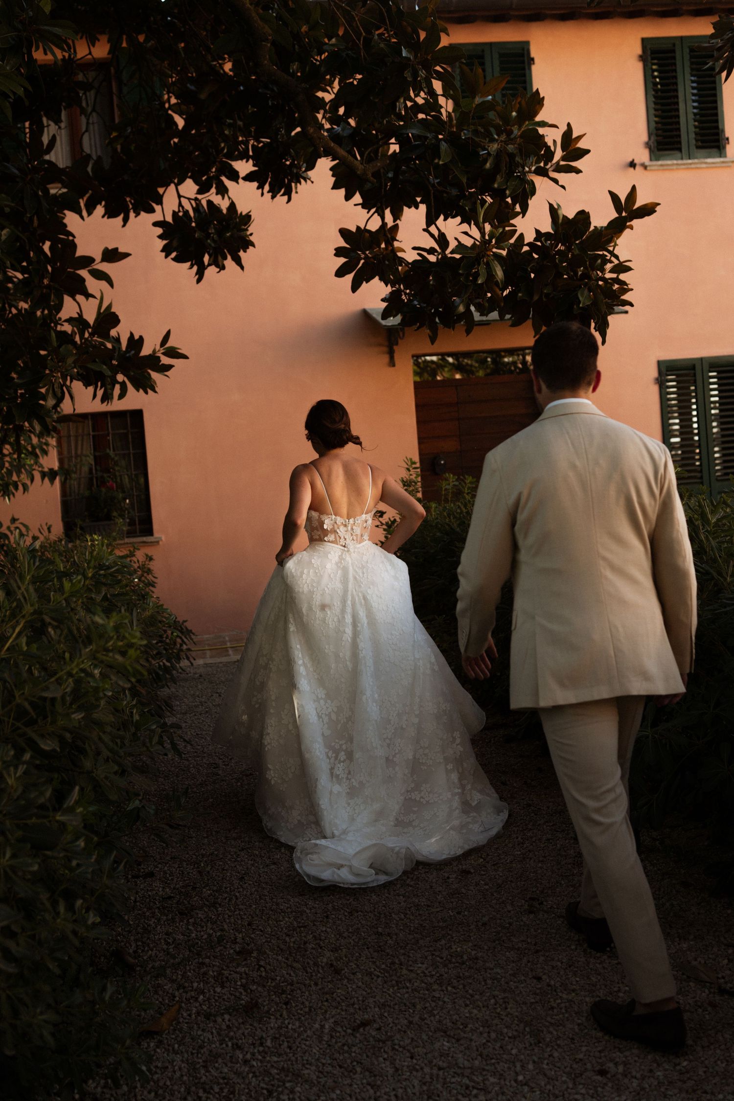 Bride in lace gown and groom in beige suit walk toward pink villa entrance surrounded by lush greenery.