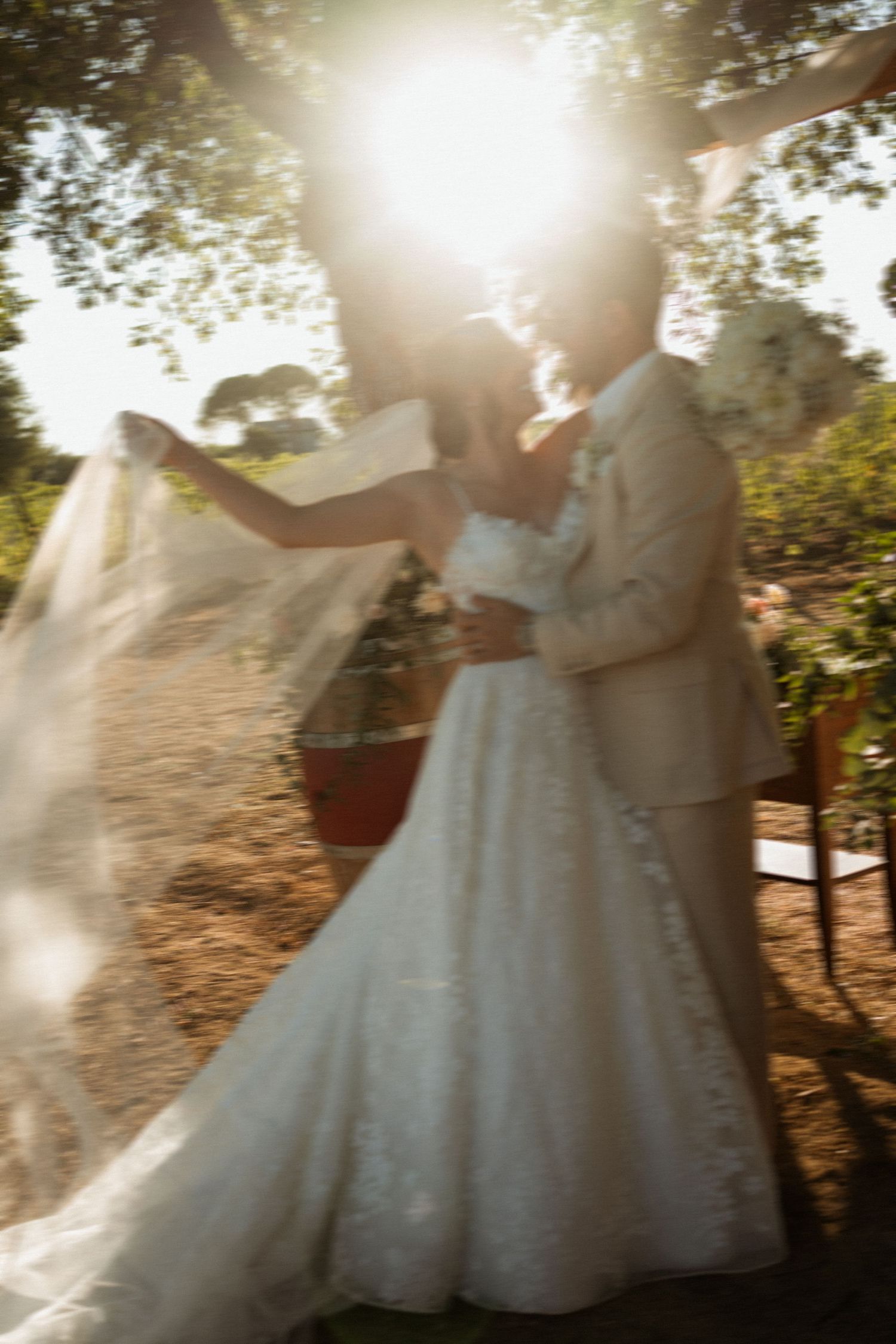 Bride and groom share romantic kiss outdoors with golden sunlight and flowing veil in dreamy bokeh effect.