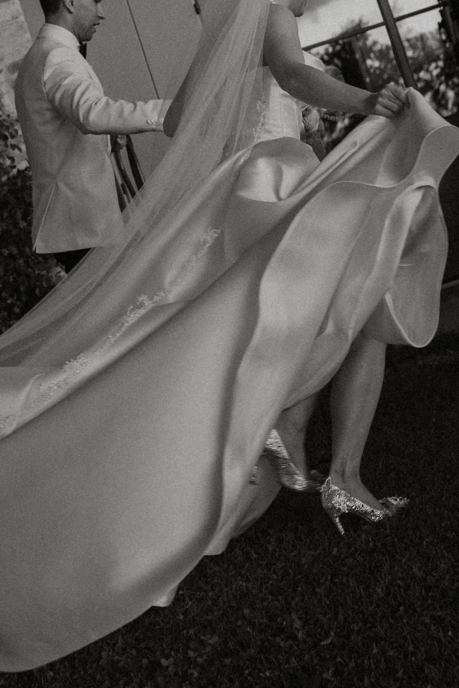 Black and white close-up of a bride's flowing wedding gown and sparkling heels on grass.
