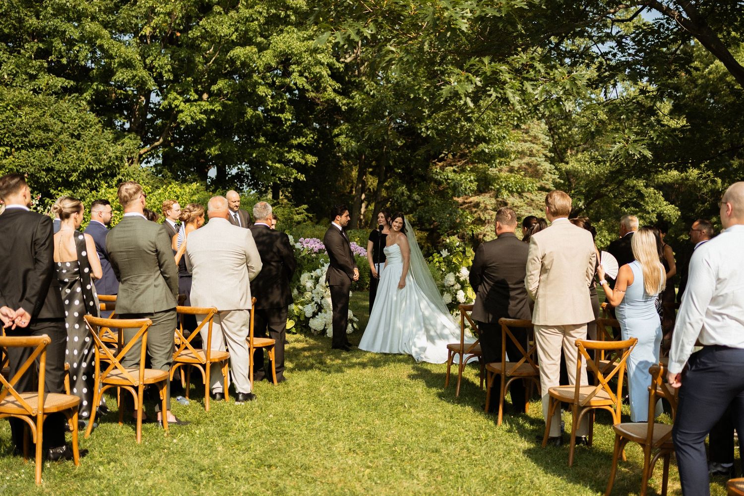 Outdoor wedding ceremony on lush green lawn with guests seated in wooden chairs facing bride and groom beneath large trees.