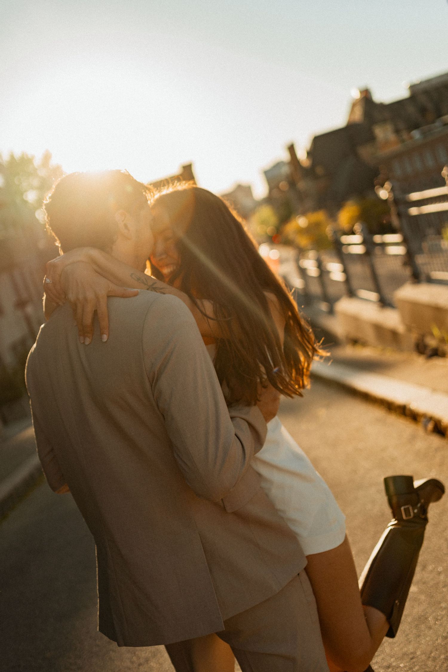 Couple sharing a romantic kiss at golden hour sunset on a city street, backlit by warm glowing sunlight.