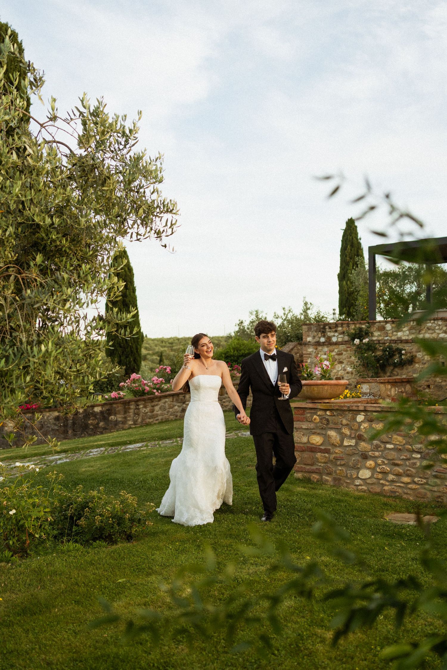Bride and groom walk hand-in-hand through lush garden grounds, bride in white gown, groom in black tuxedo holding champagne.