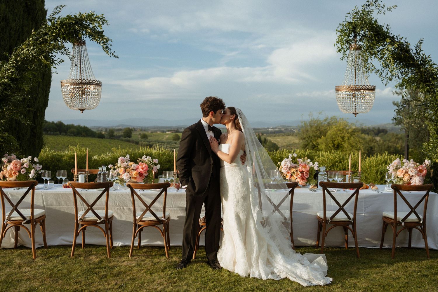 Bride and groom kissing at outdoor Tuscany wedding reception with chandeliers, floral arrangements, and rolling hills backdrop.