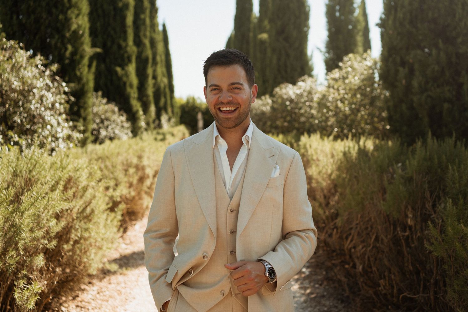 Smiling man in beige linen suit standing on garden path surrounded by cypress trees and lush greenery.