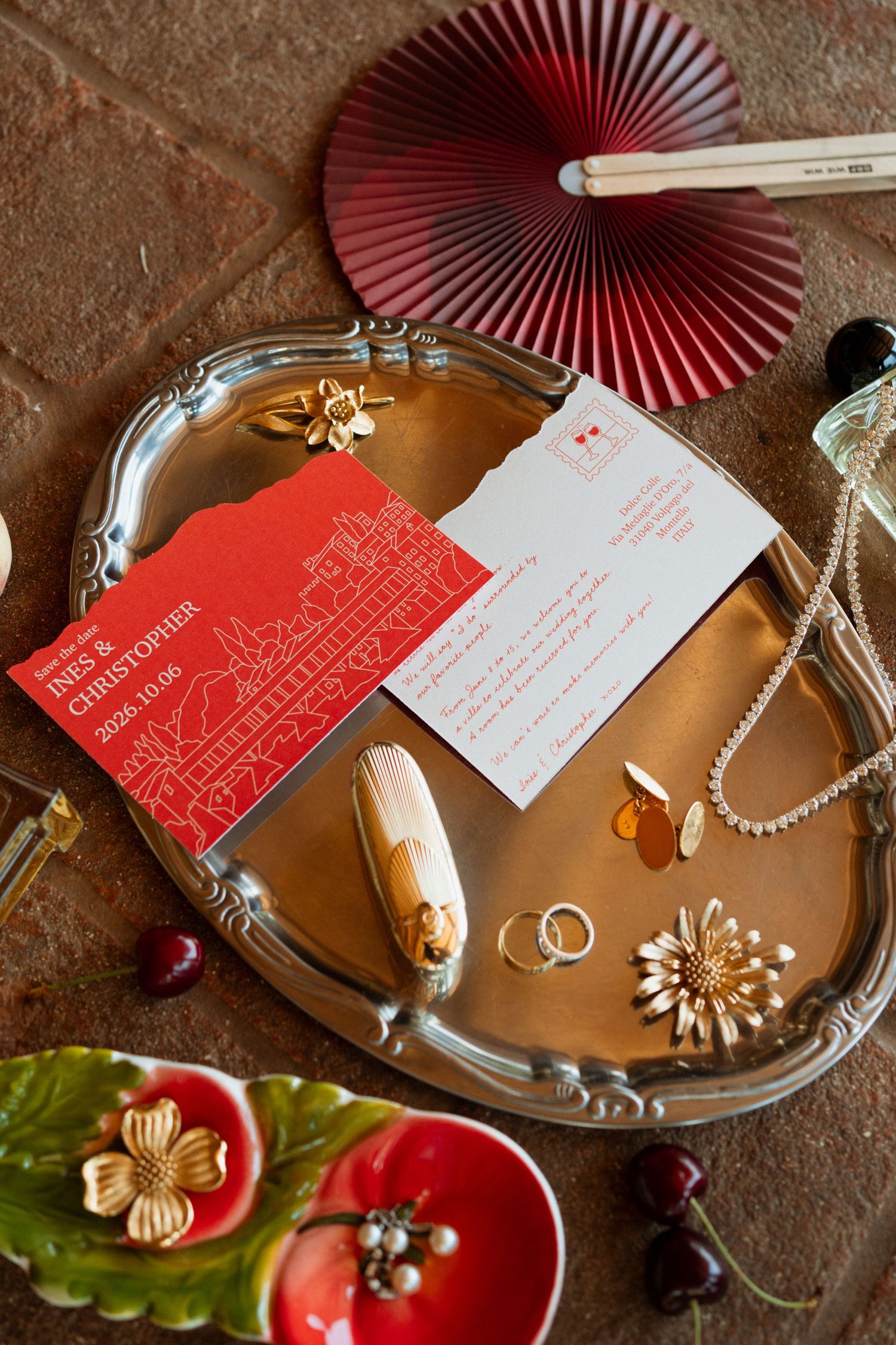 Silver tray with wedding invitations, gold jewelry, and rings beside a red paper fan on a brick surface.