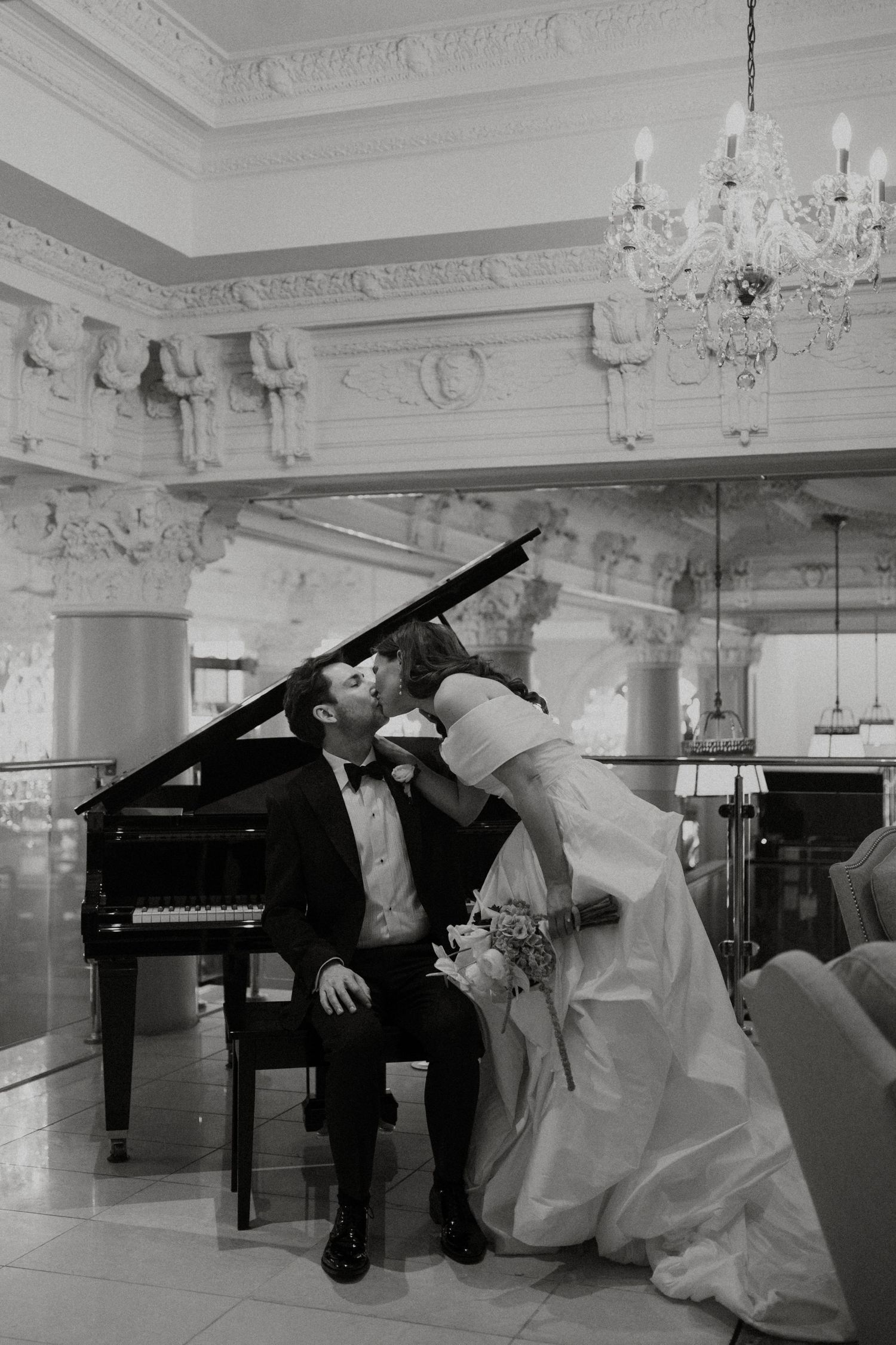 Bride and groom share romantic kiss at grand piano in ornate ballroom with crystal chandelier.