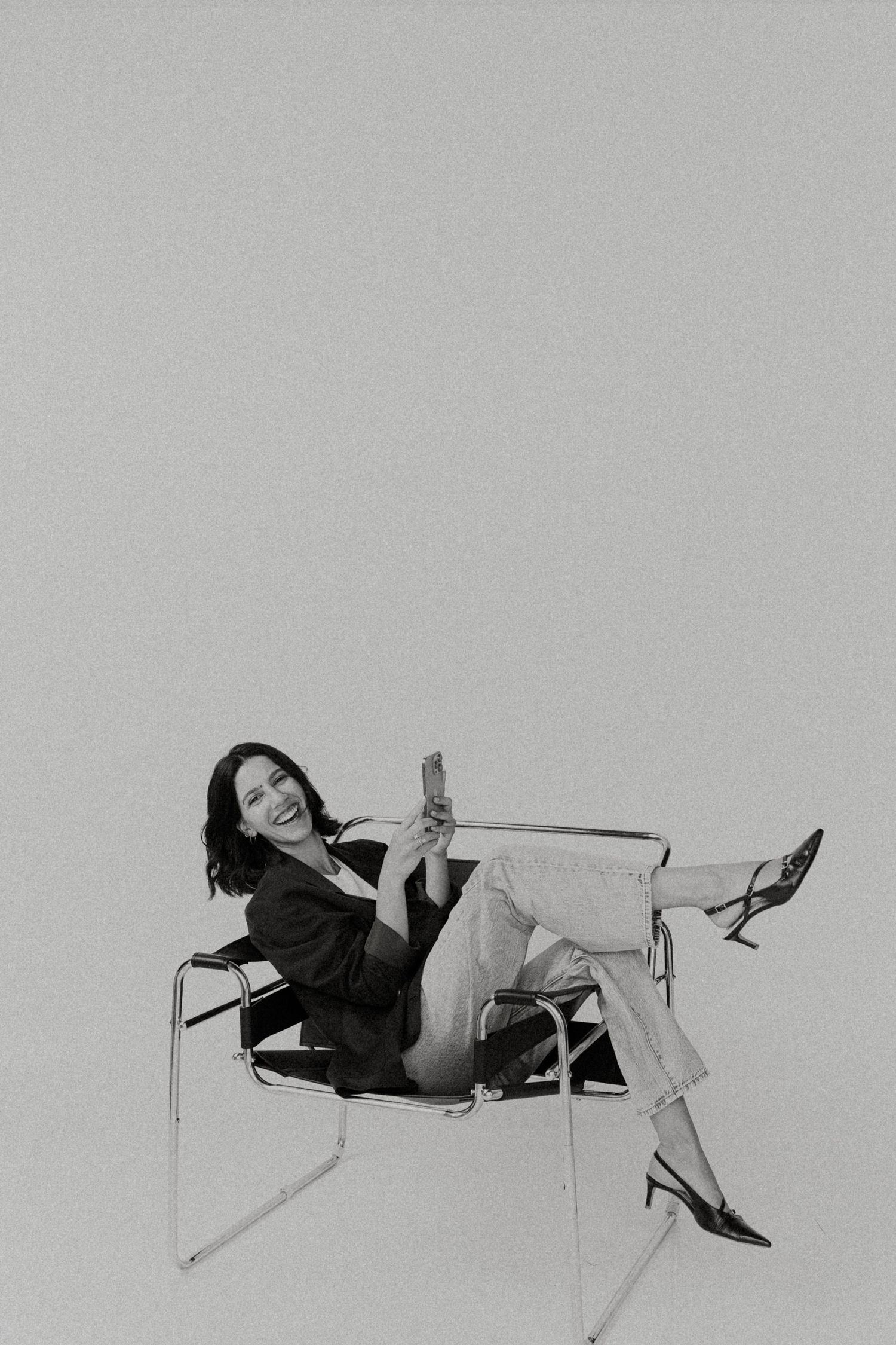 A woman in a black blazer and jeans sits relaxed in a Wassily chair holding a phone in a black and white photo.