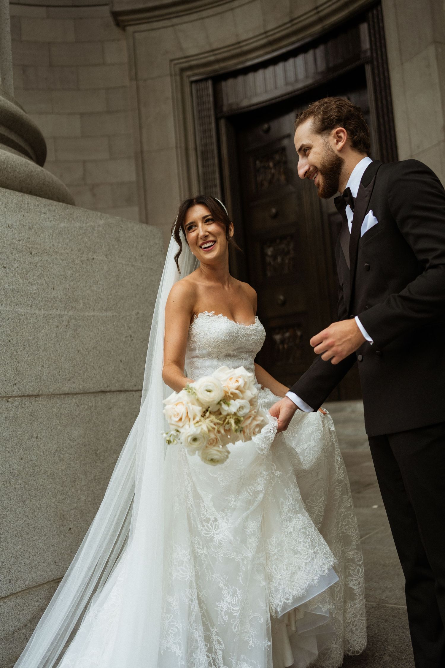 Smiling bride in strapless lace gown with veil and groom in tuxedo laughing on stone steps.
