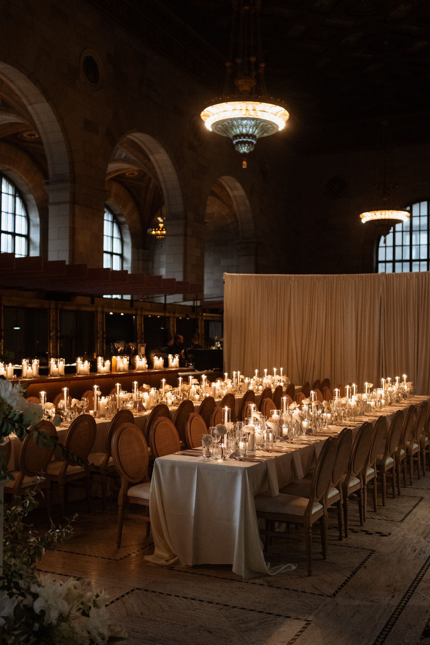 Grand wedding reception hall with long candlelit tables, ornate chandeliers, and arched windows.