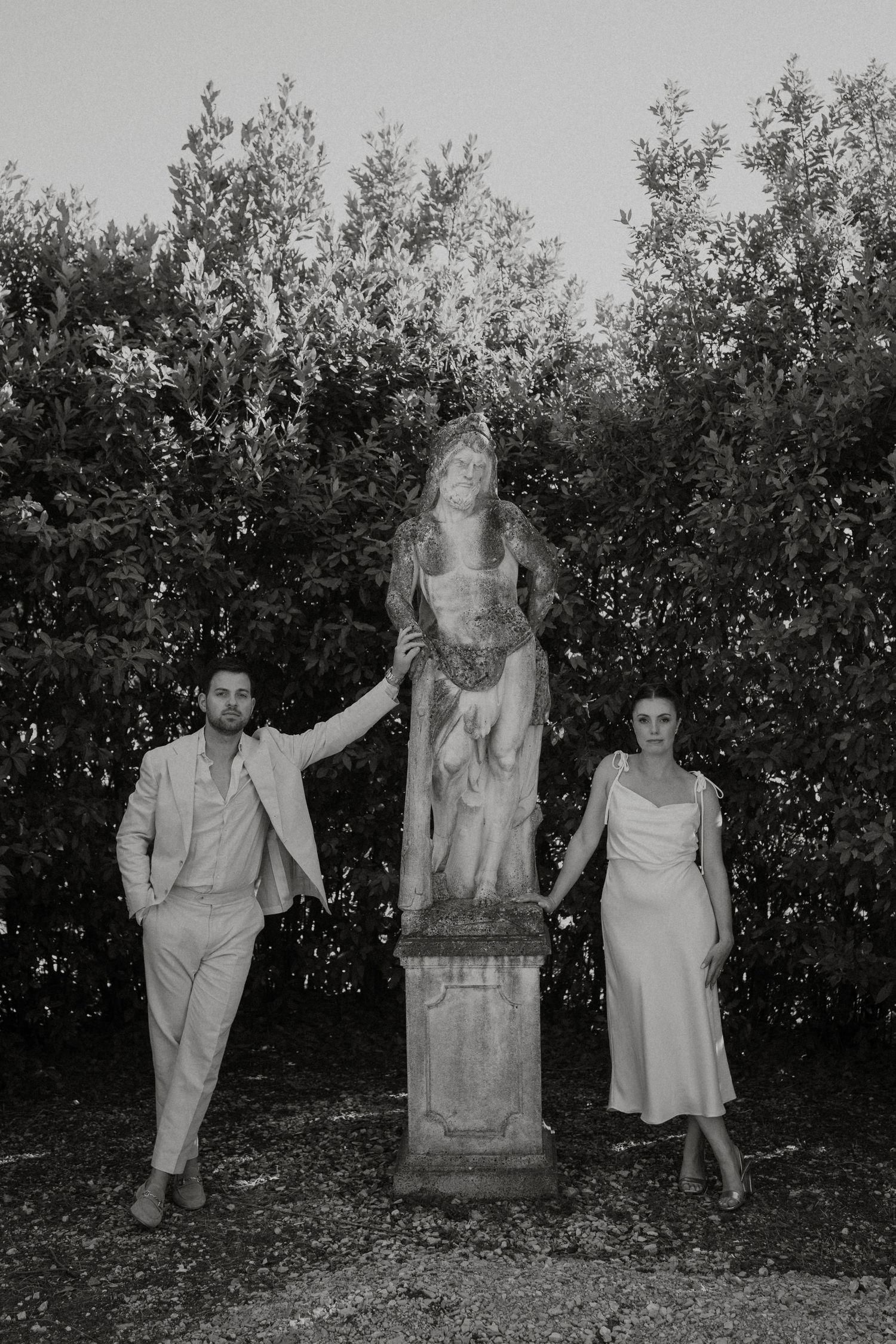Black and white photo of a couple posing beside a classical stone statue in a lush garden setting.