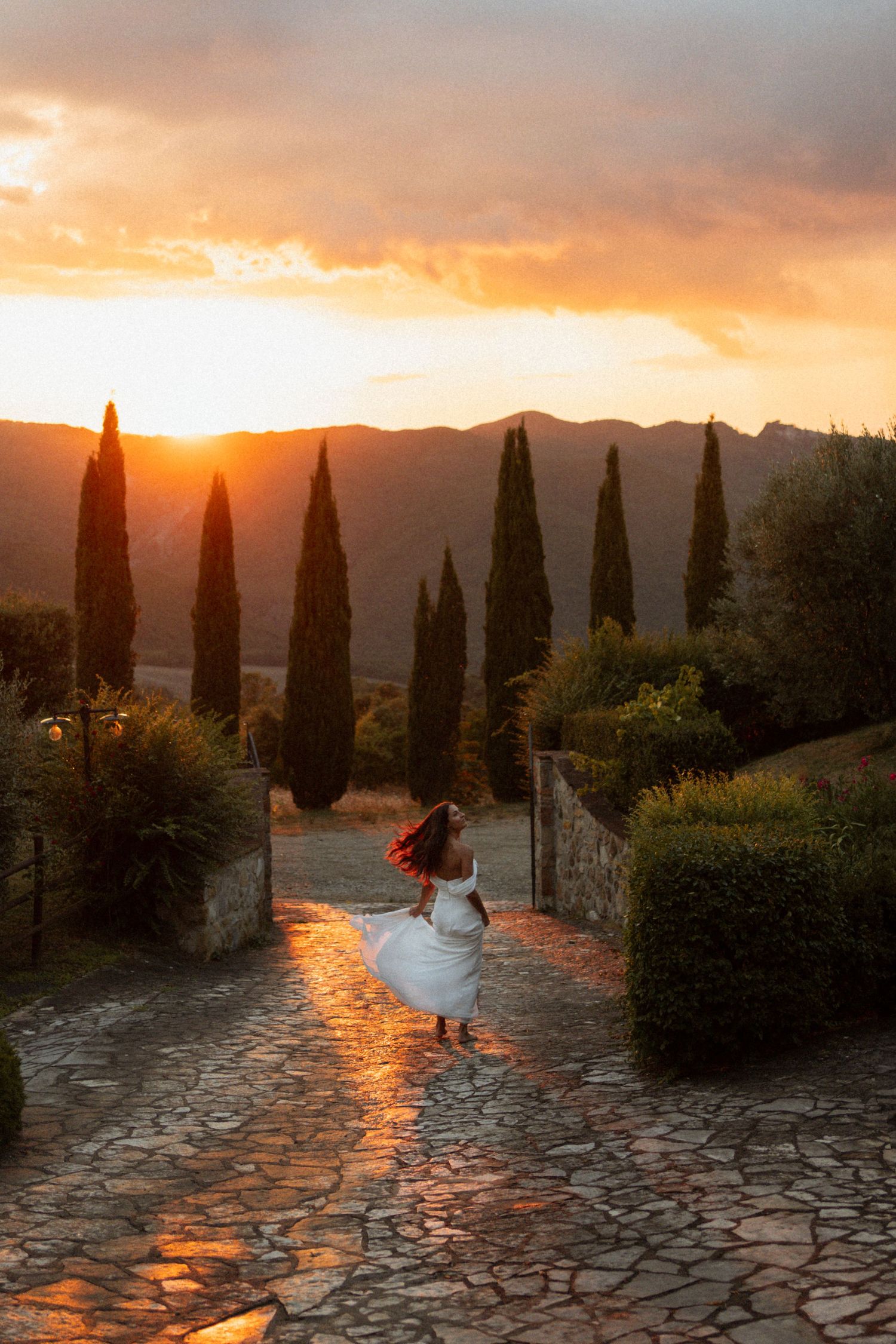 Bride in white dress twirling on stone pathway at golden sunset with cypress trees and mountains in Tuscany.