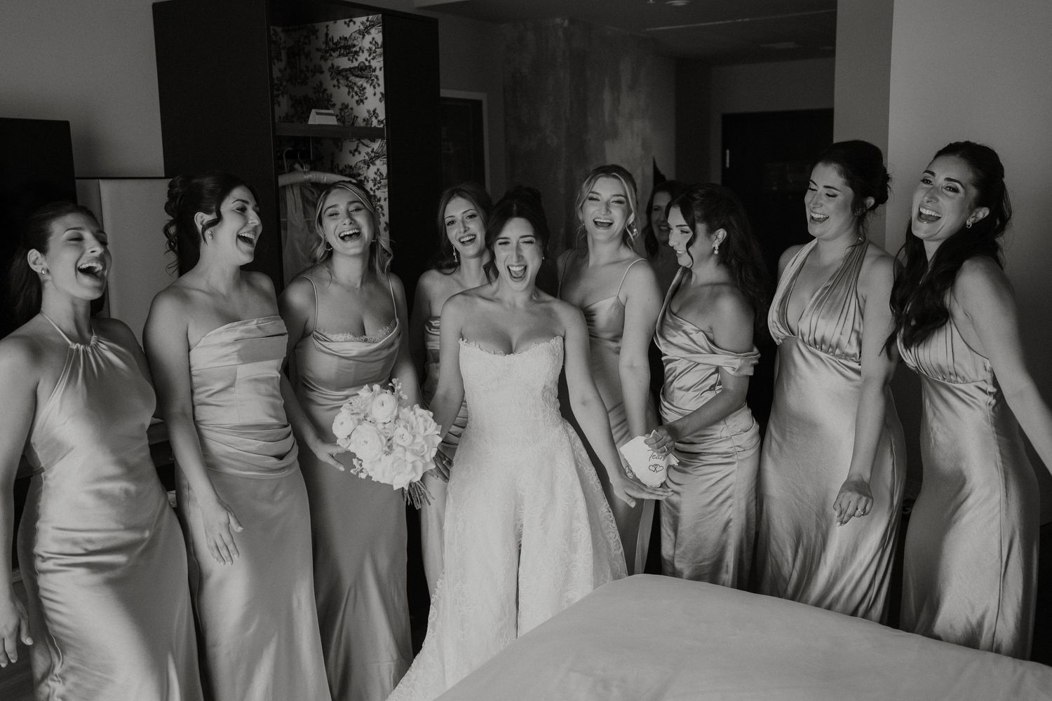 Black and white photo of bride with bridesmaids in satin gowns laughing together.