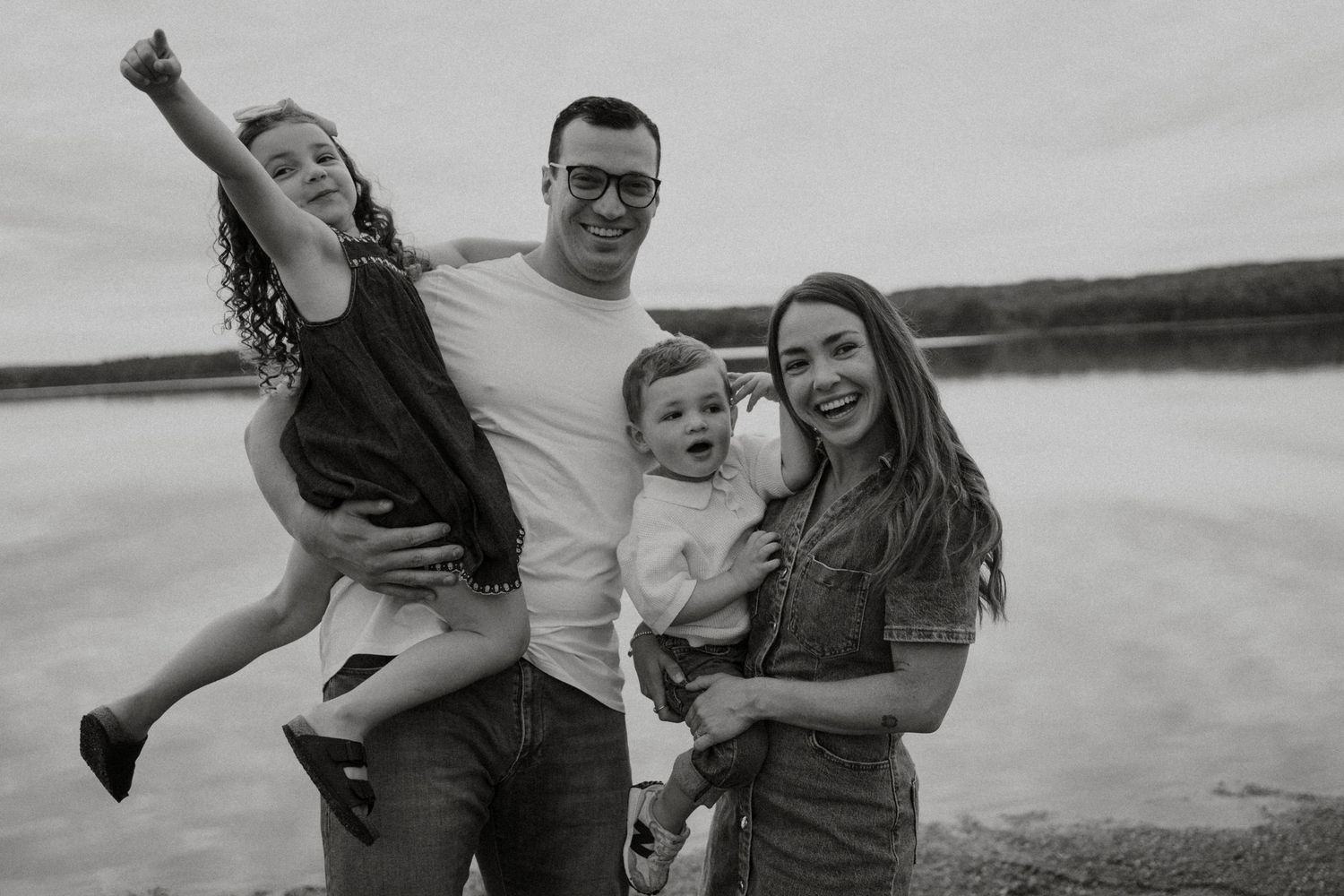 Black and white family photo of parents with two young children laughing by a lake shore.