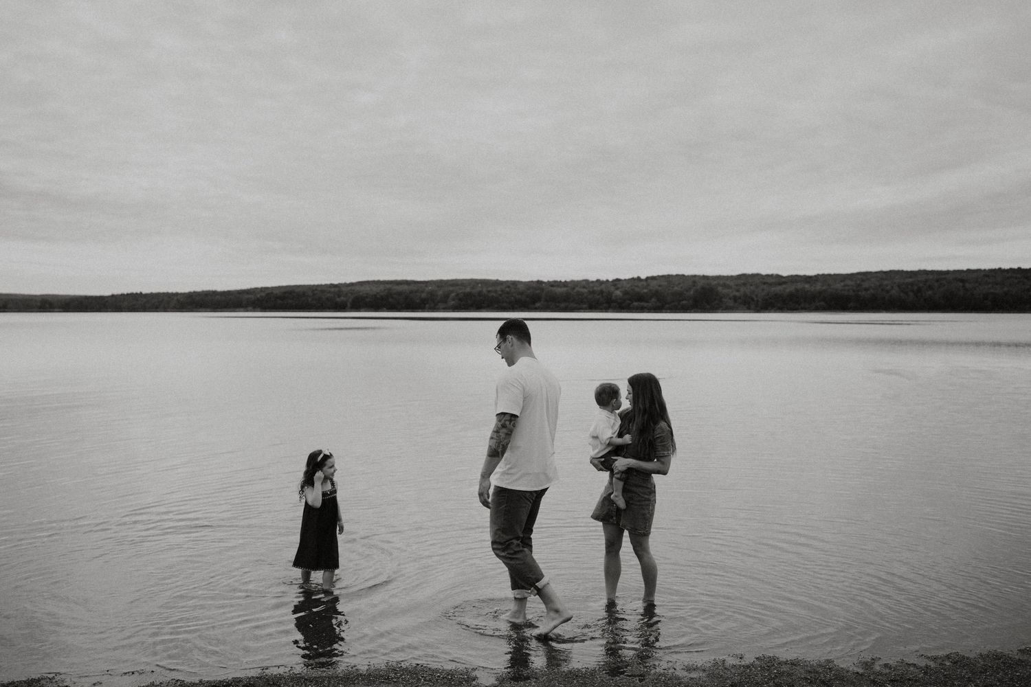 A family of four wades in a calm lake in a serene black and white photo.