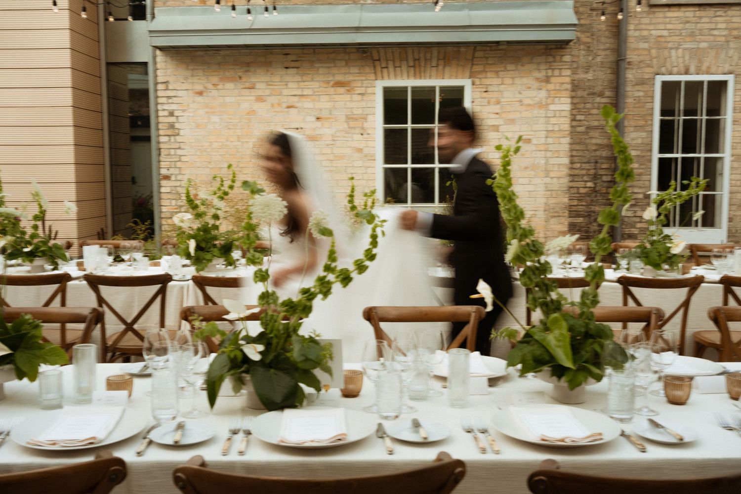 Blurred bride and groom dance beside elegantly set outdoor wedding reception table with lush greenery centerpieces.