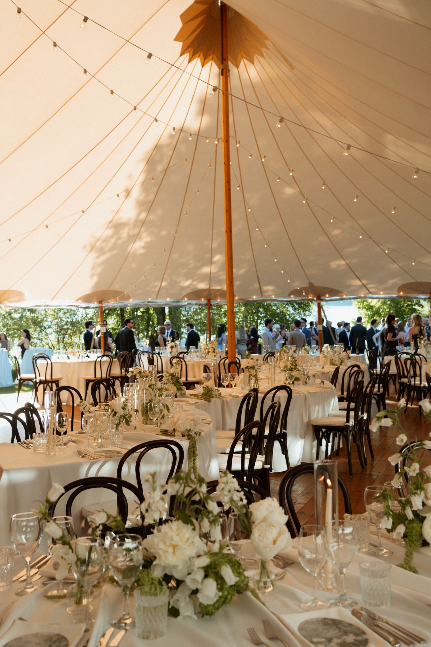Elegant wedding reception under a sailcloth tent with white floral centerpieces, taper candles, and bentwood chairs on round tables.