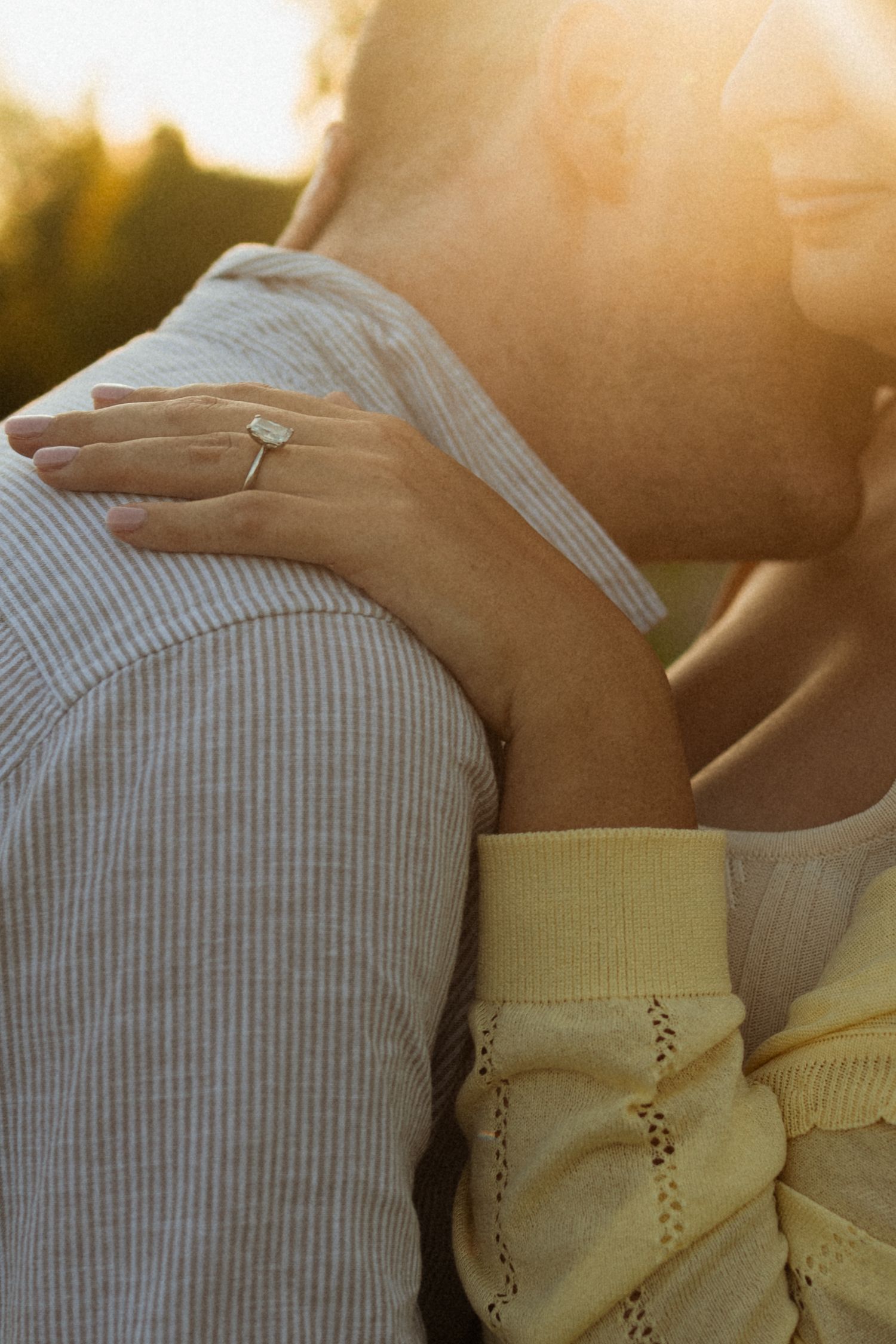 Couple embracing at golden hour sunset, woman wearing elegant oval engagement ring on hand resting on mans shoulder.