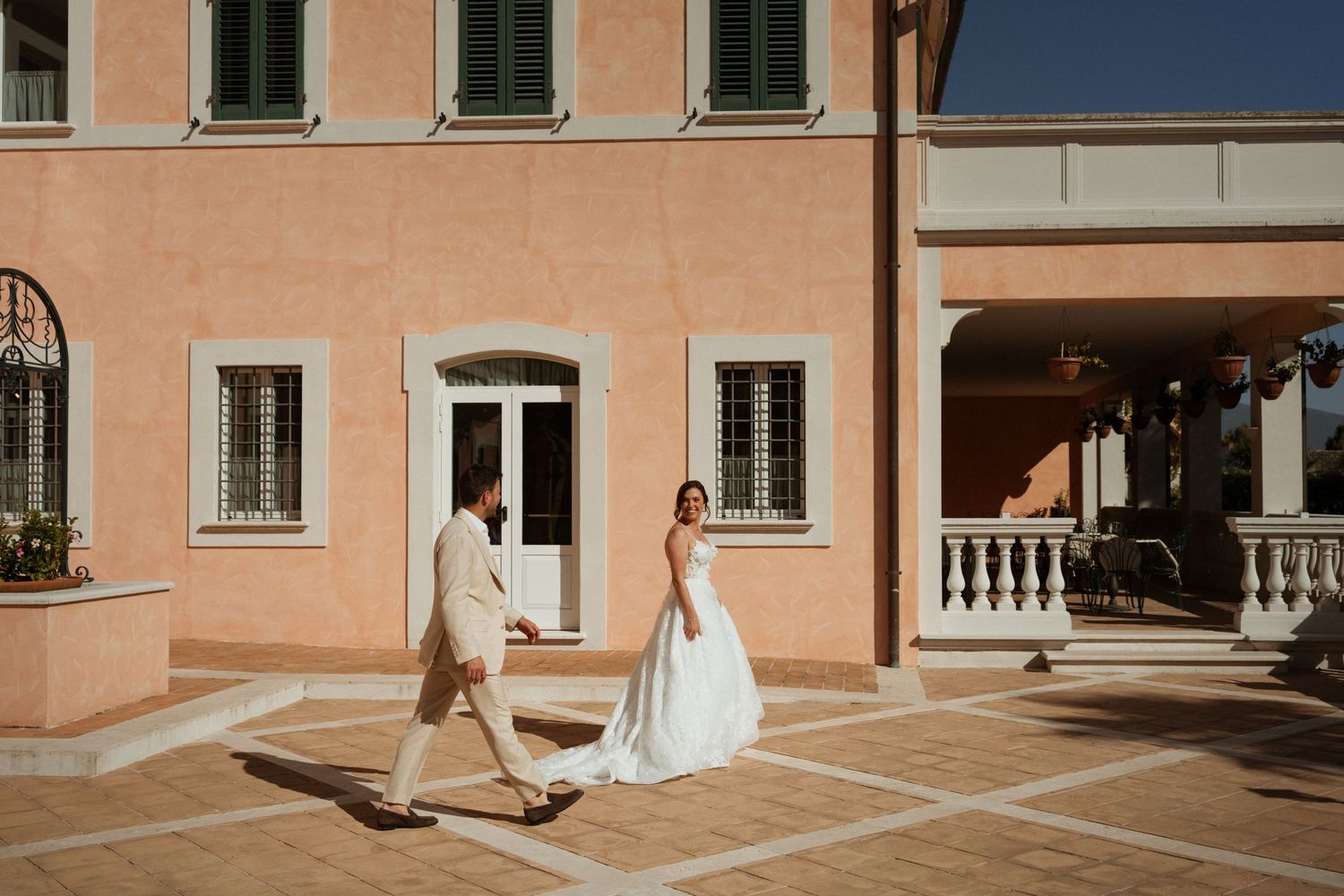 Bride and groom walk hand in hand across a sunlit courtyard beside a elegant pink Italian villa.