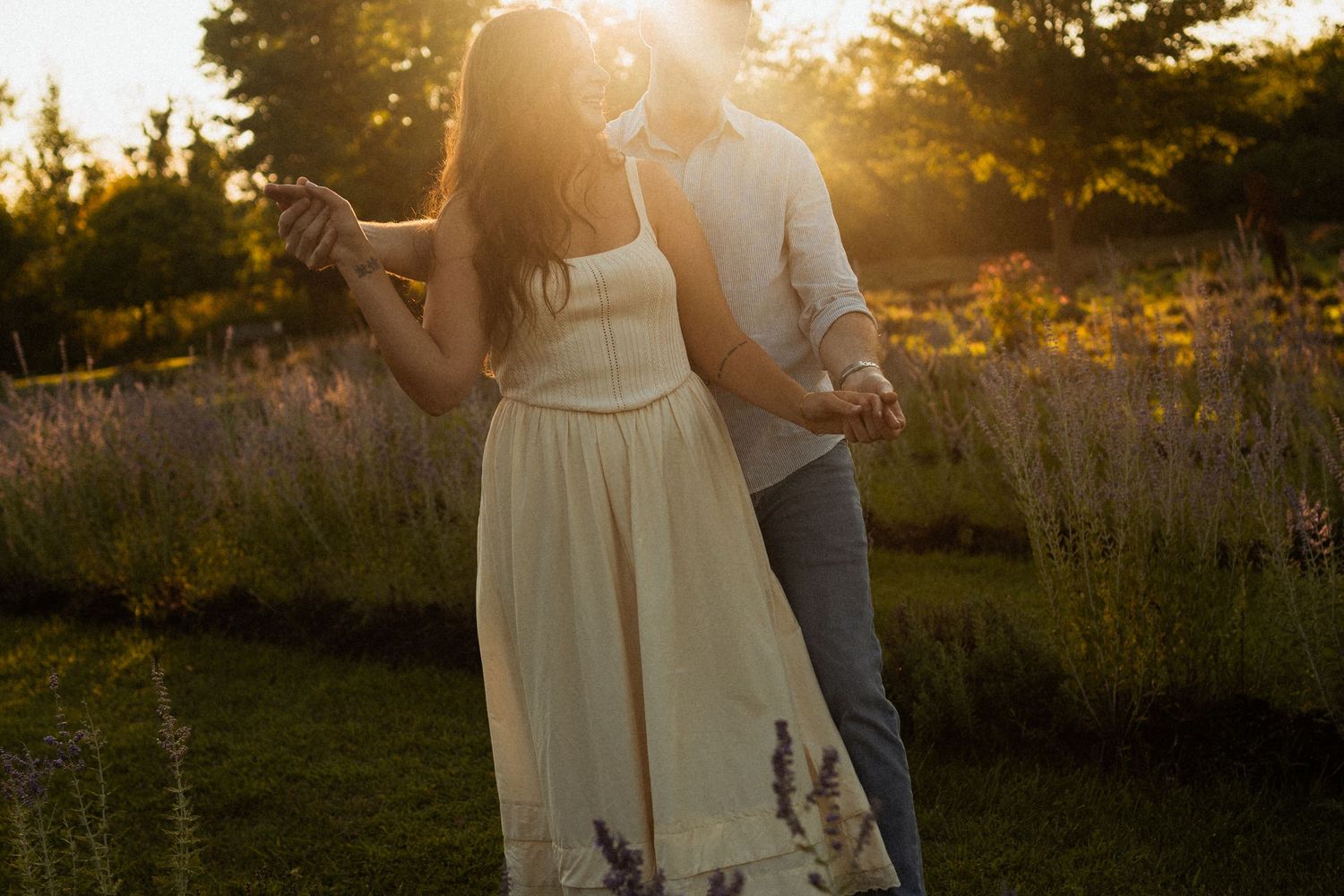 Couple dancing in golden sunset light surrounded by lavender fields, woman in flowing cream dress holding hands with man.