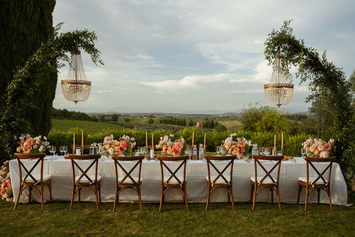 Elegant outdoor wedding reception table with floral arrangements, chandeliers, and vineyard views in Tuscany.