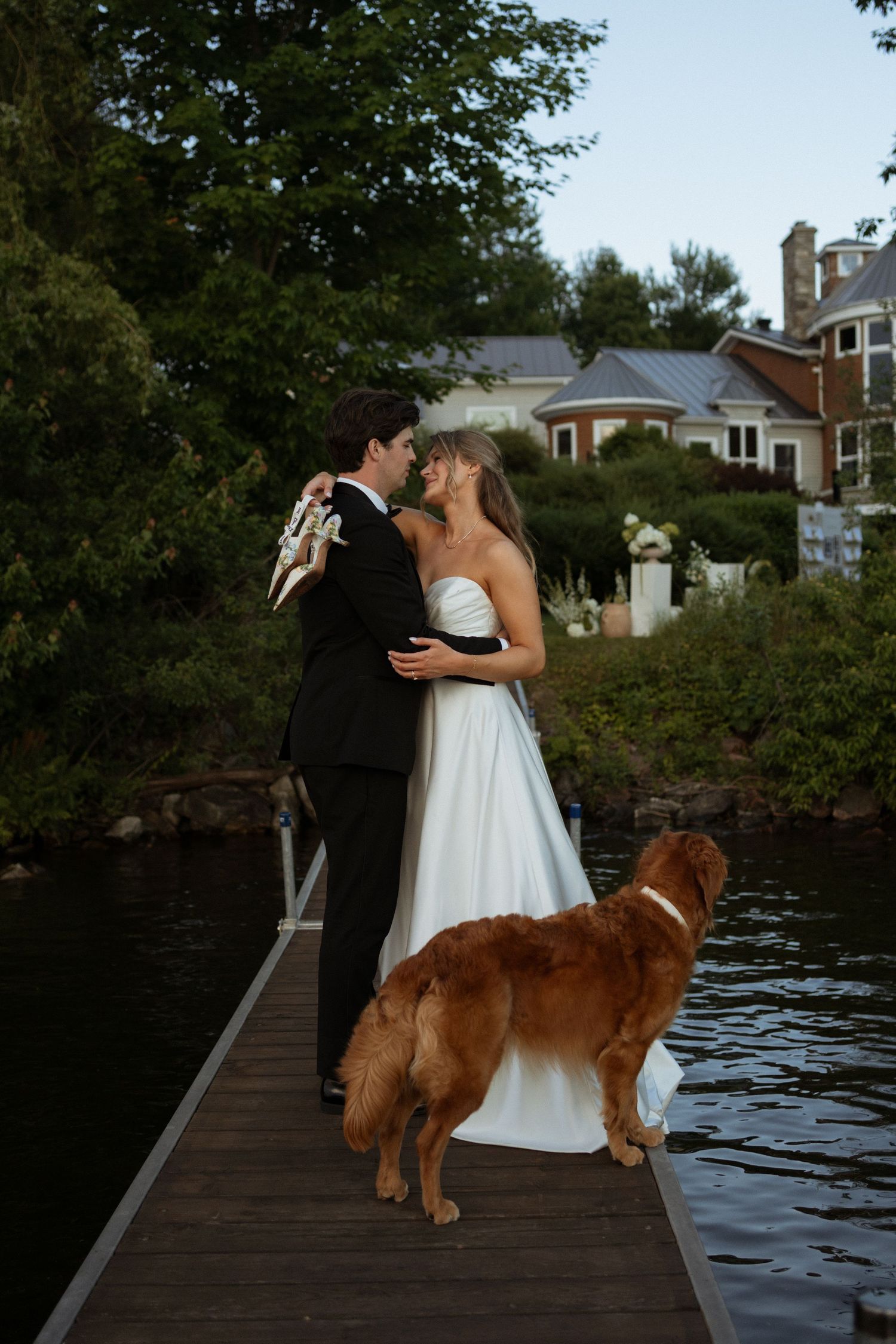 Bride and groom kissing on lakeside dock at dusk with golden retriever dog standing beside them.