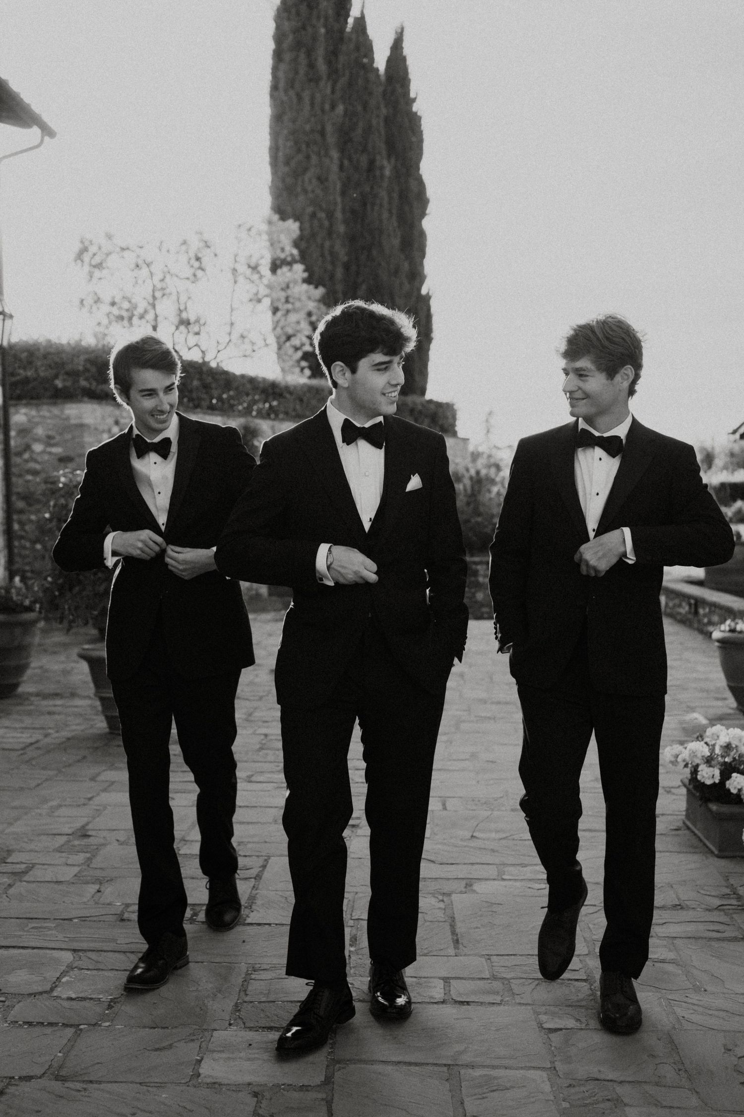 Three groomsmen in black tuxedos and bow ties walking outdoors in black and white photo.