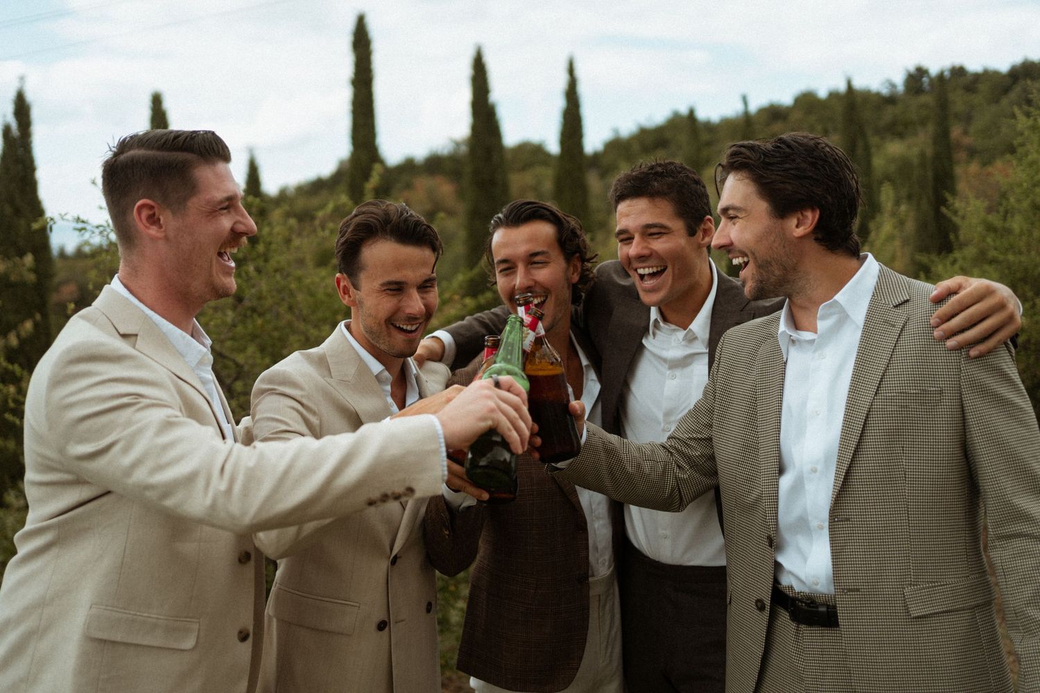 Group of groomsmen in suits toasting with beer bottles outdoors in Tuscany.