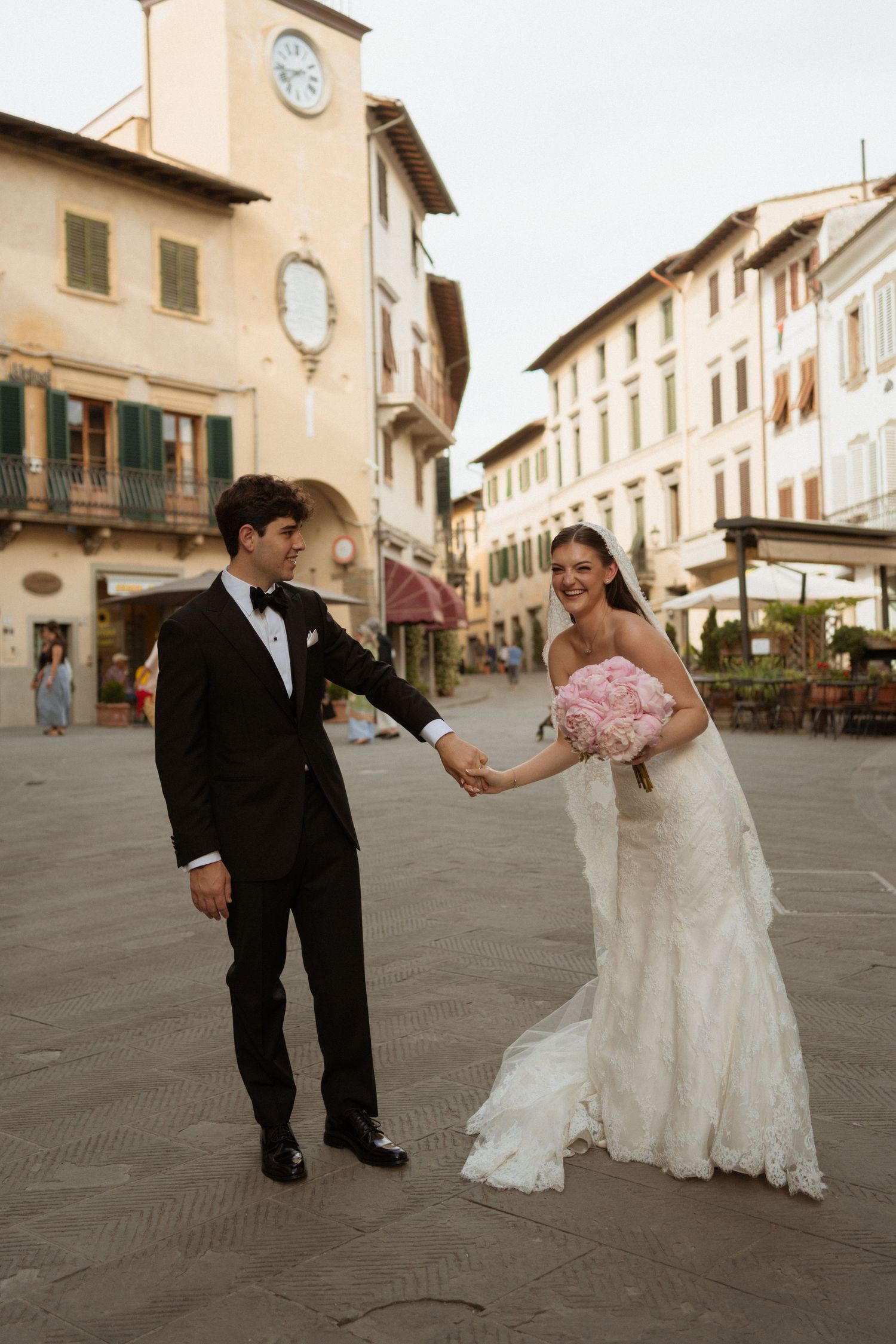 Bride and groom holding hands in Italian piazza with clock tower backdrop in Tuscany.