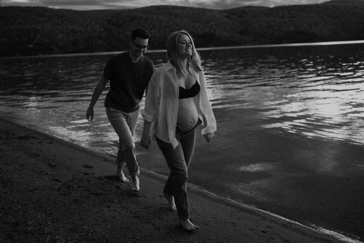 Black and white photo of a pregnant couple walking along a lakeside beach at dusk, woman in bikini top.