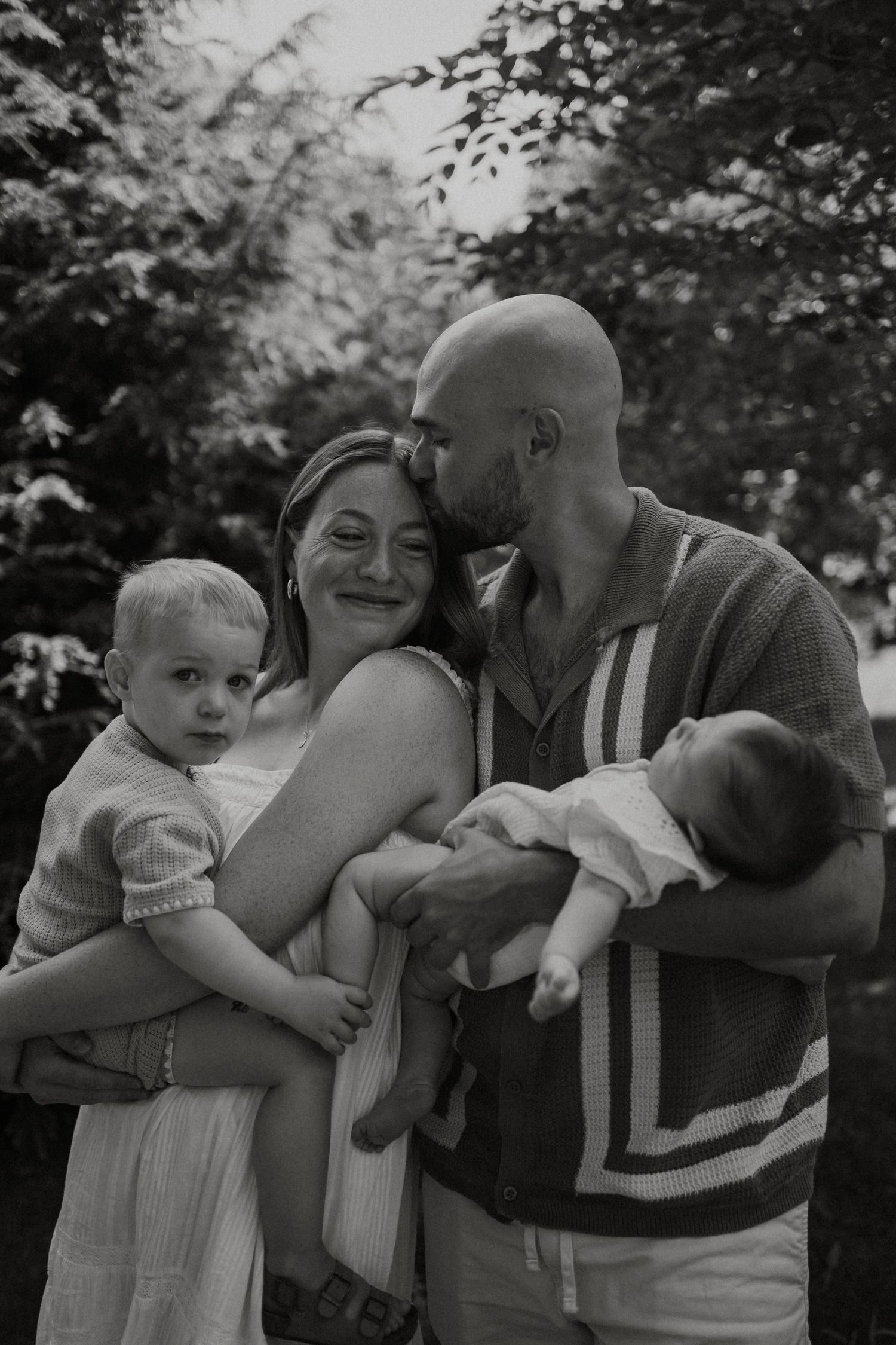 Black and white family portrait of parents holding two young children outdoors, sharing a tender moment.