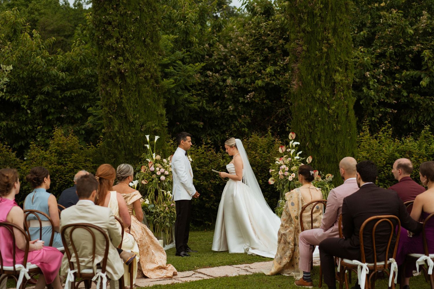 Outdoor wedding ceremony with bride and groom at floral altar, surrounded by lush greenery and seated guests.