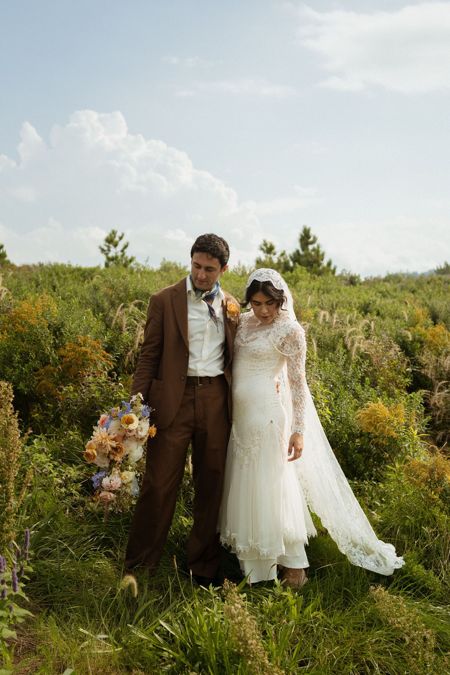 A bride in white lace gown and groom in brown suit stand together in a lush green field holding a colorful bouquet.