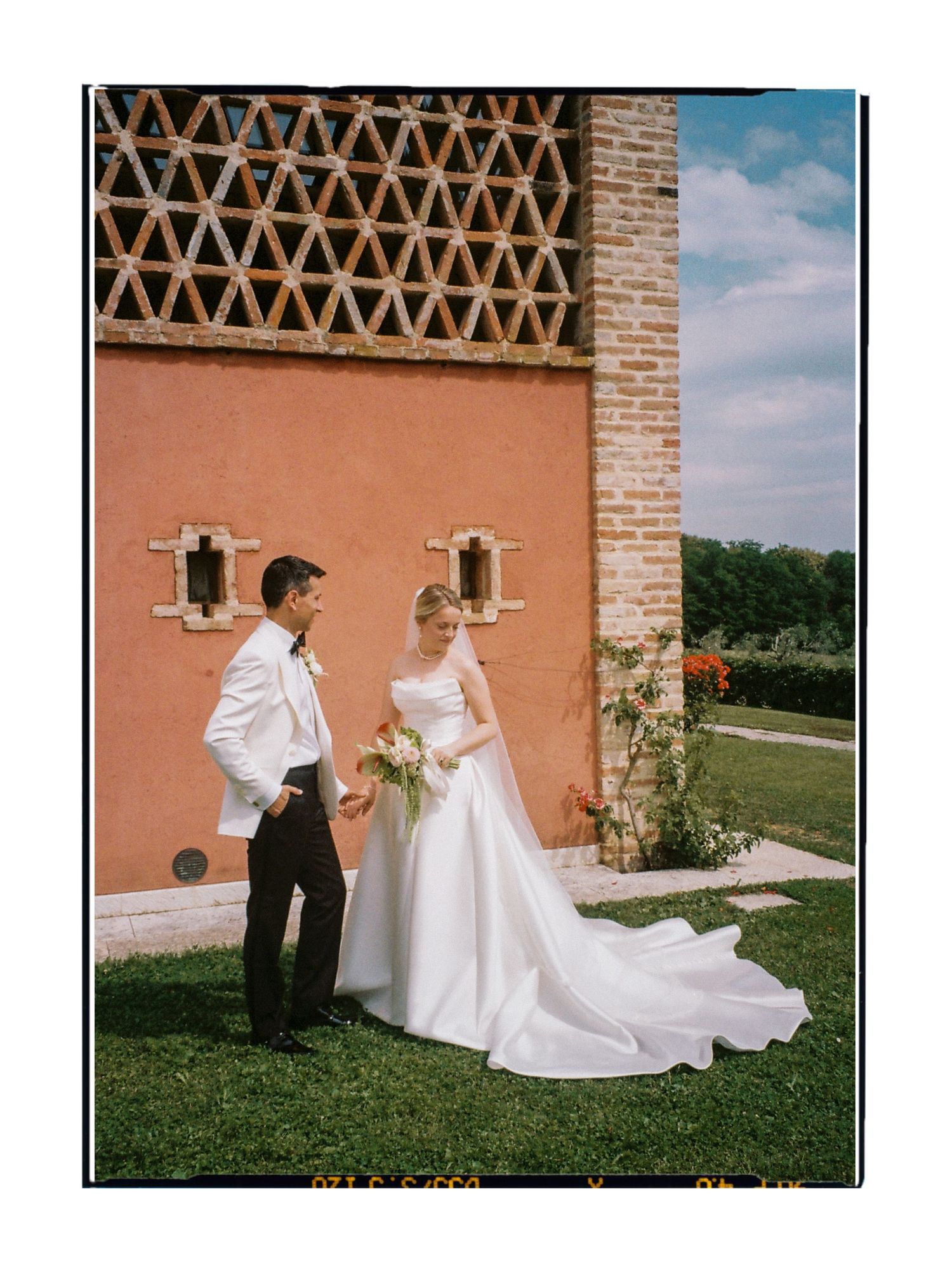 A bride and groom pose outside a rustic terracotta villa with lush gardens on their wedding day.