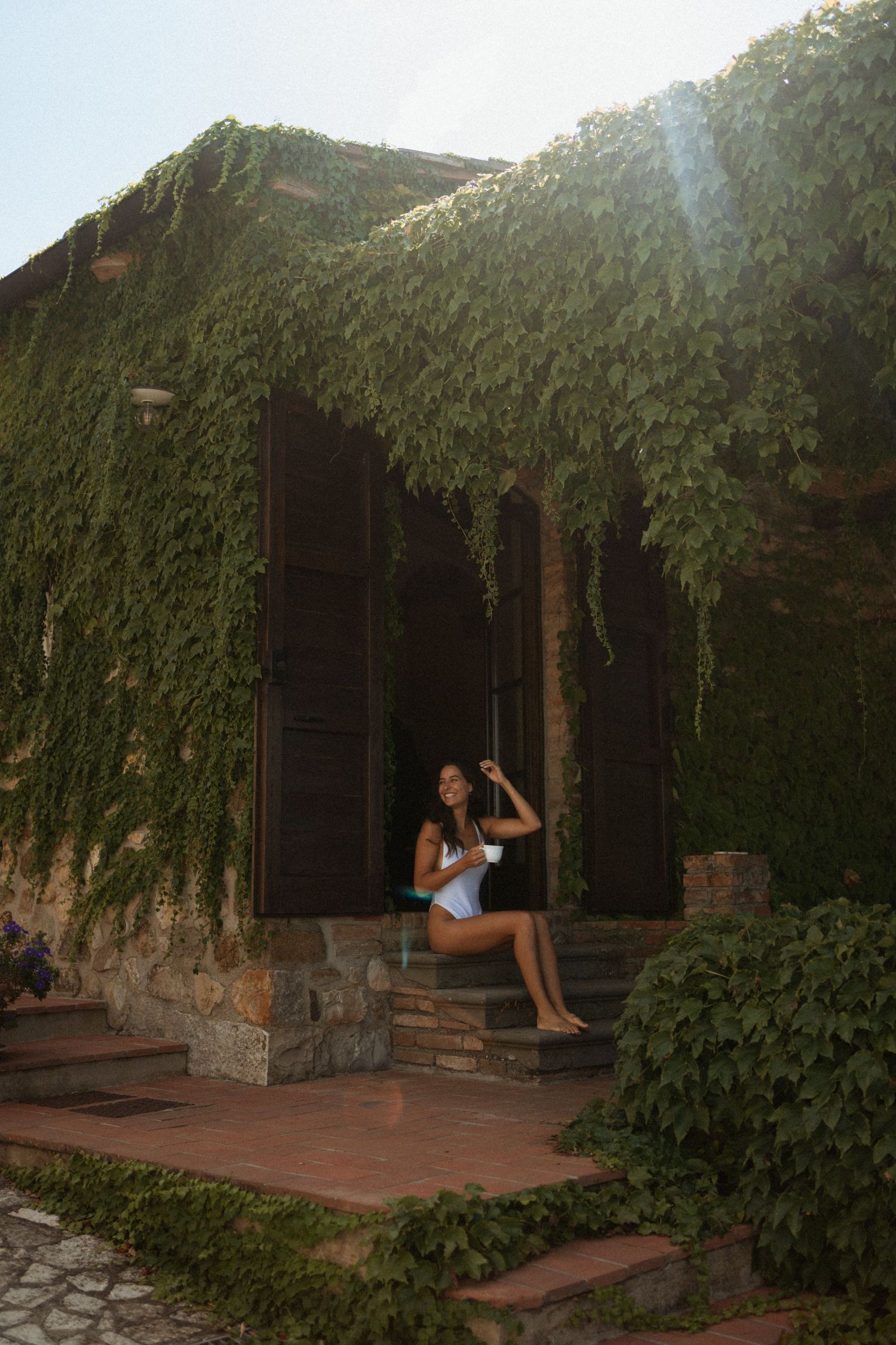 Bride in white mini dress sitting on ivy-covered stone villa steps in Tuscany.