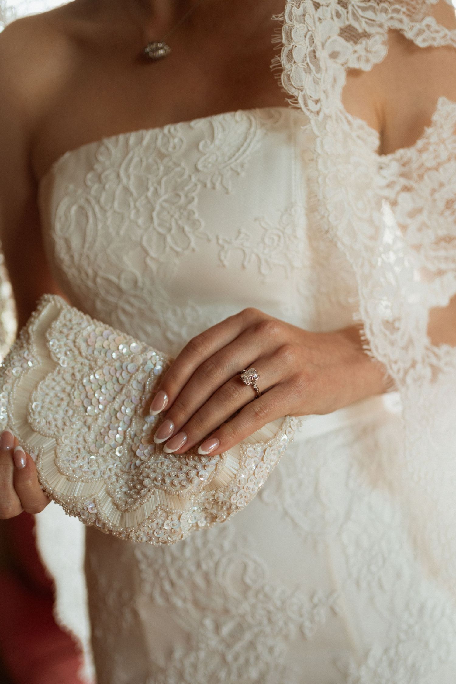 Bride in strapless lace wedding gown holding beaded clutch with diamond engagement ring.